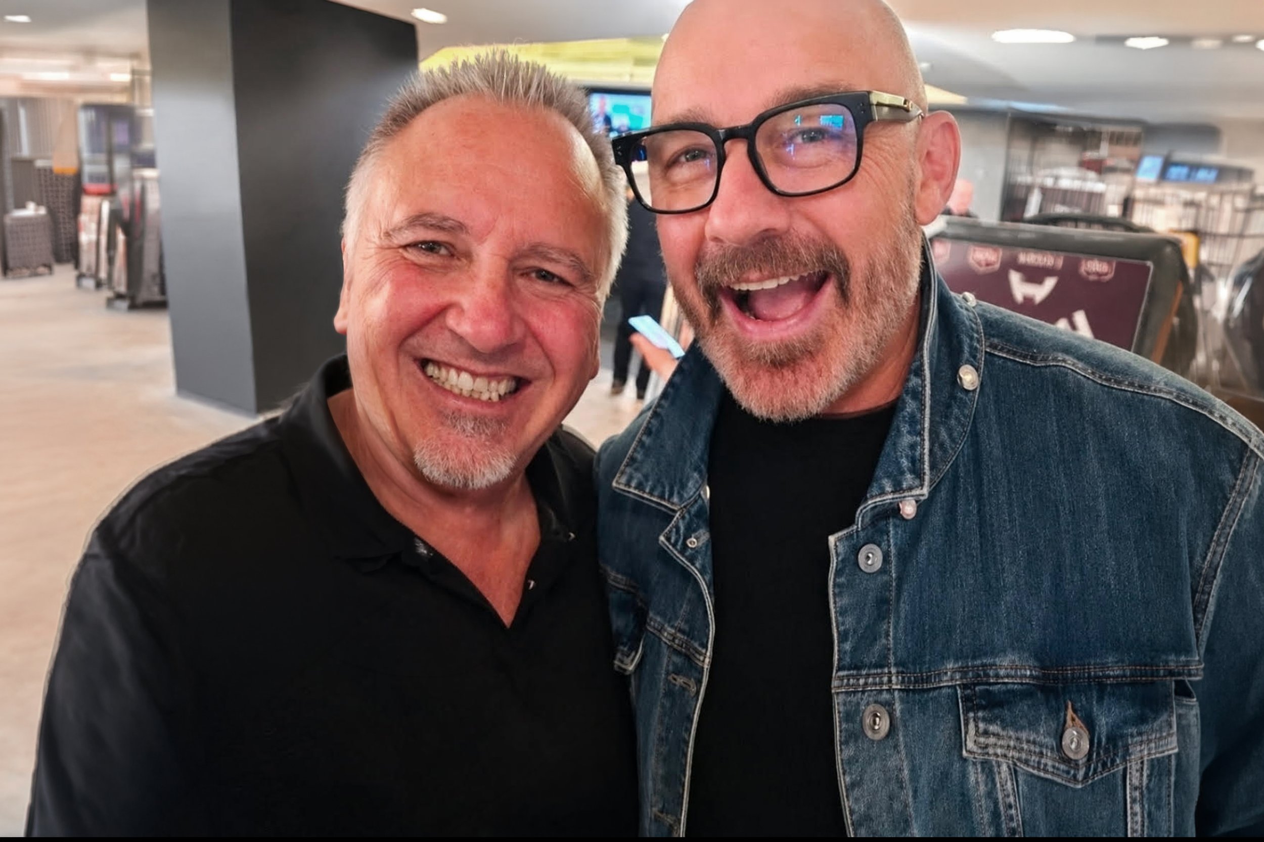Two smiling men taking a selfie in an airport, with baggage and airport signage in the background.
