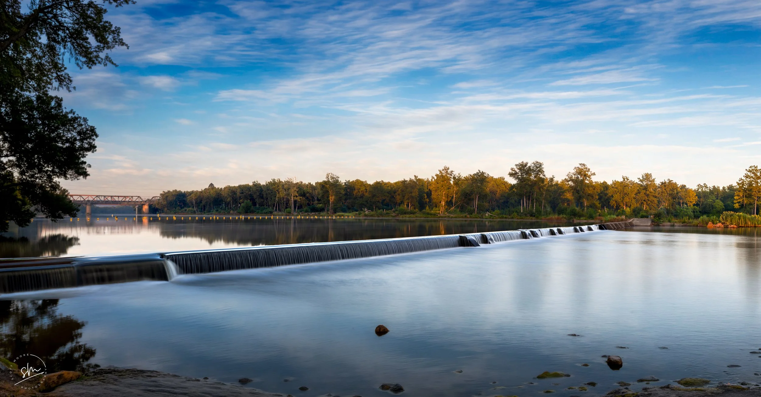 A peaceful river with a small waterfall, surrounded by trees under a blue sky with scattered clouds.
