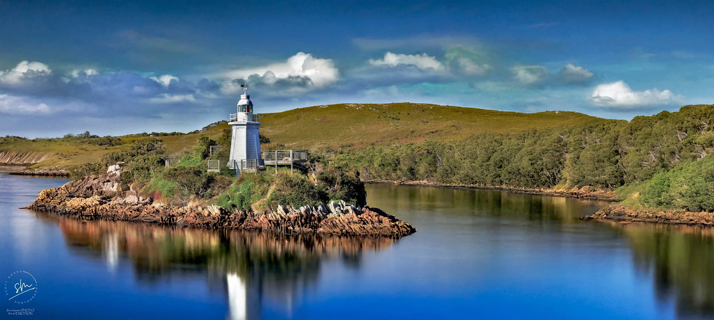 A white lighthouse on a small rocky island surrounded by calm water and lush green hills under a partly cloudy sky.