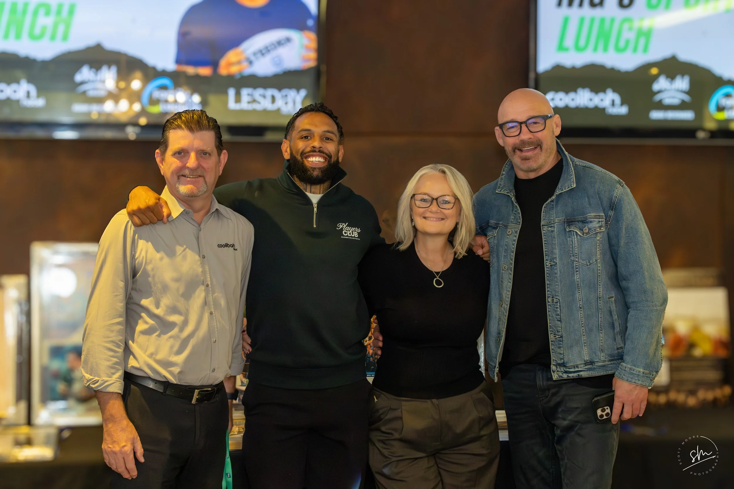 Four smiling adults standing together indoors, with two large screens displaying the words "Lunch" and "Tuesday" in the background. Coolibah Hotel Merrylands, The Fox and Mark Geyer with Michelle Becari and the Lesday Group of Hotels Mg's big lunch