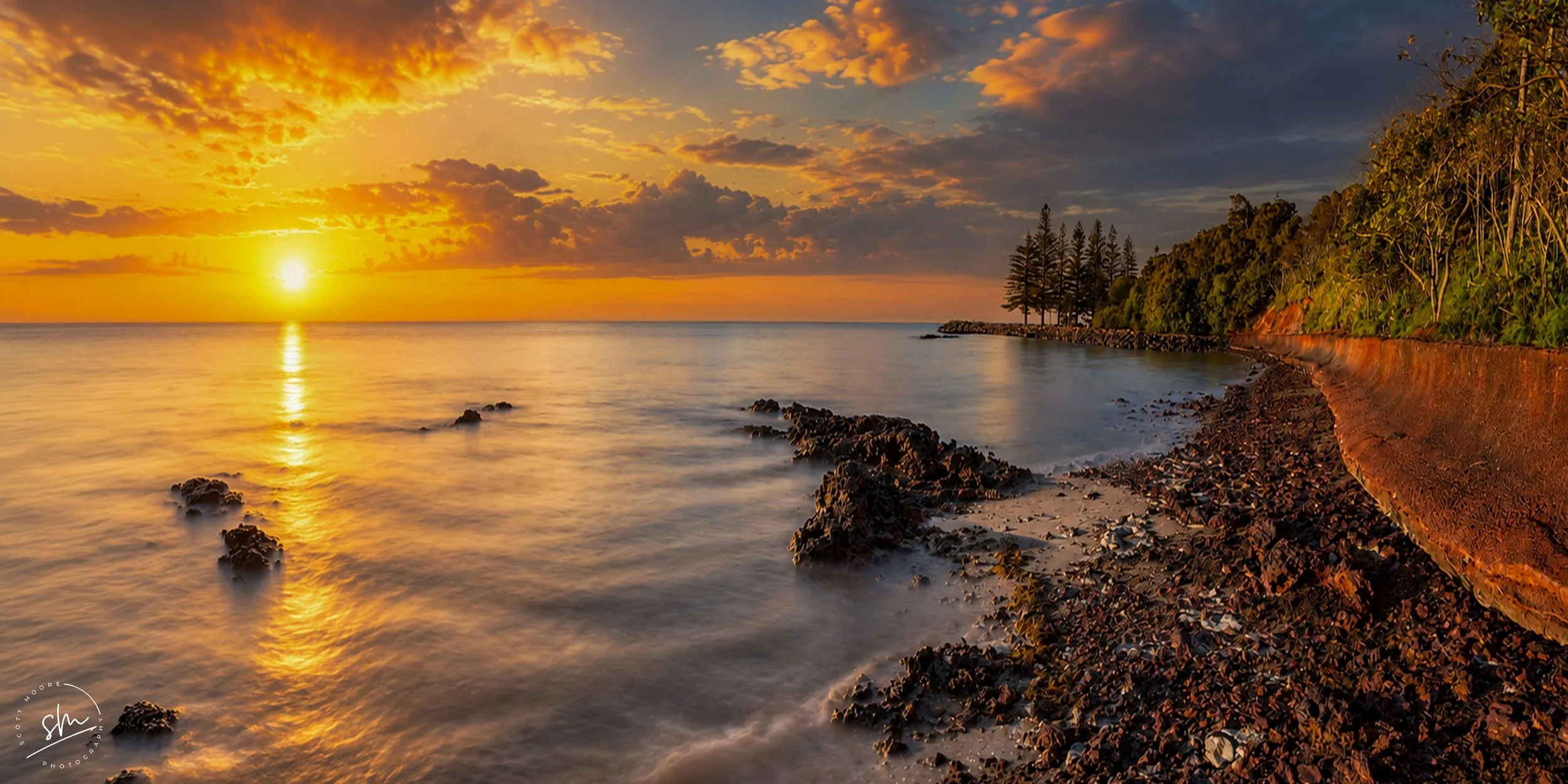 Sunset over a calm ocean with a rocky shoreline, trees on a small hill, and a partly cloudy sky with orange and yellow hues.