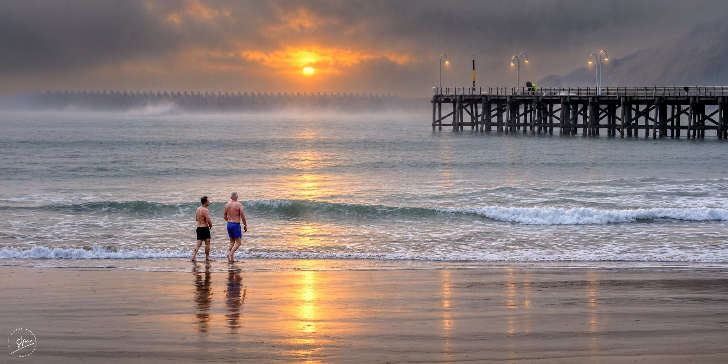 Two men walking along the shoreline at sunset or sunrise with a pier extending into the water and a foggy mountain in the background.