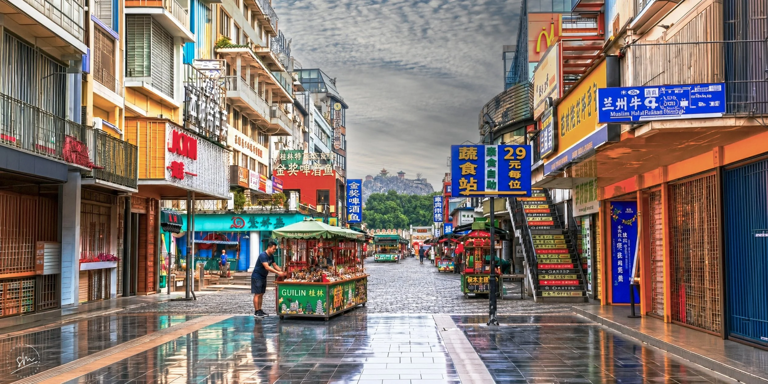 A vibrant street scene with colorful signage in Chinese and English, a small food stand labeled GUILIN, and a person browsing the items, with buildings, shops, and a historic hilltop structure in the background.