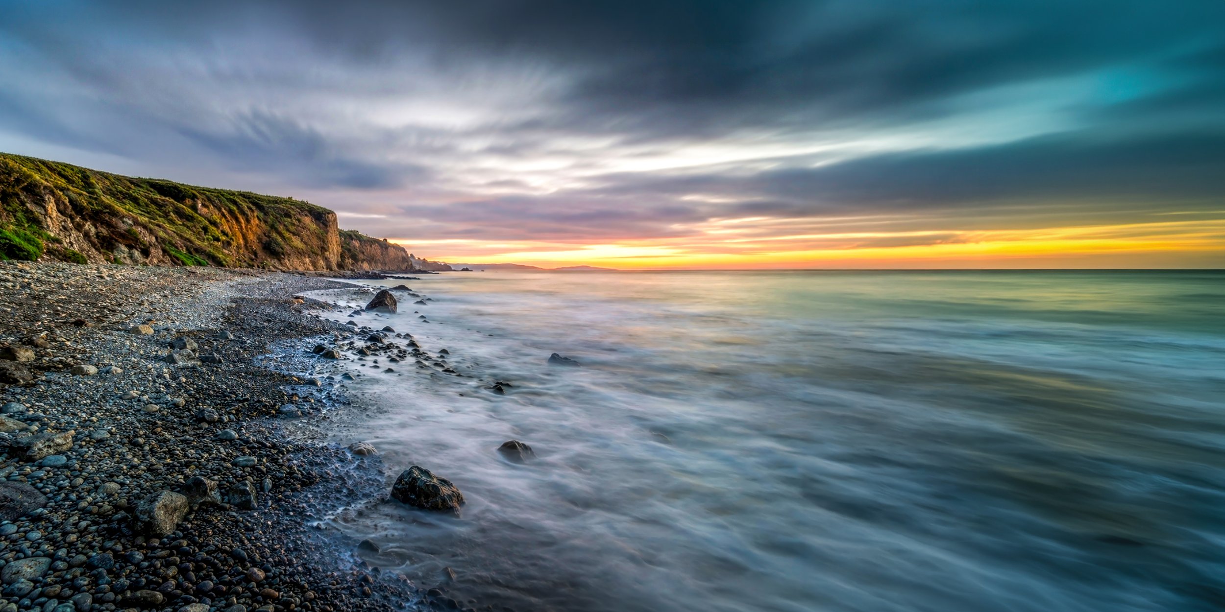 A scenic view of a rocky beach at sunset with cliffs on the left, ocean waves in the foreground, and a vibrant sky with clouds and the setting sun in the distance.