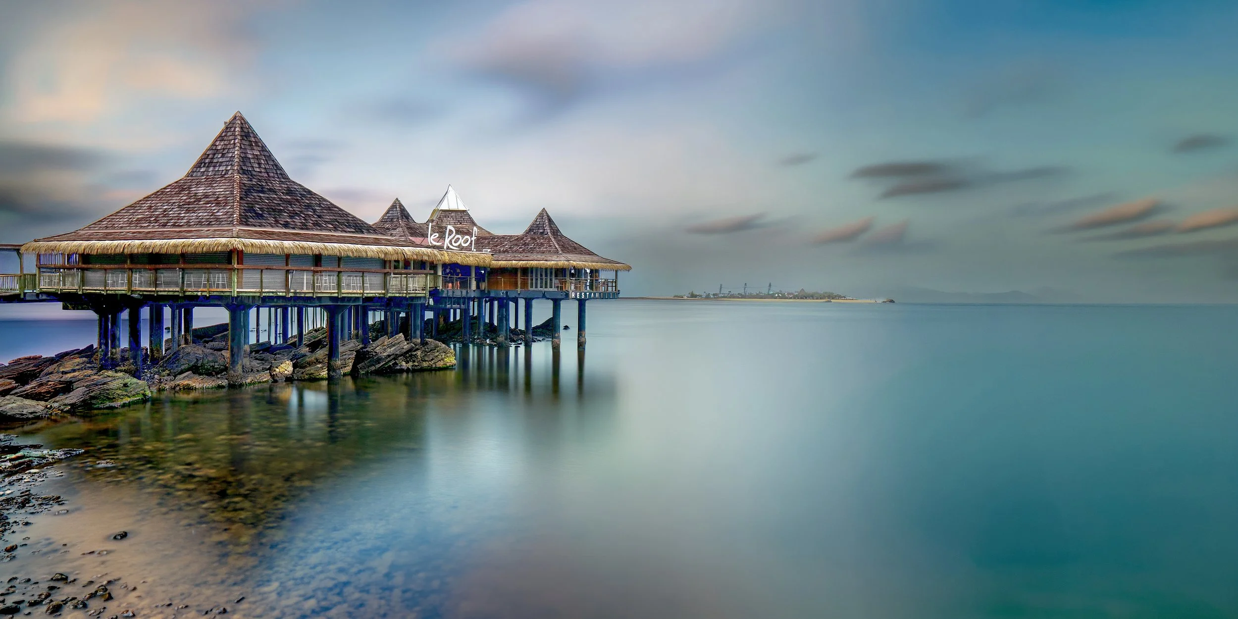 A floating restaurant with a thatched roof and wooden pillars, named le Root, positioned on rocks by calm water under a partly cloudy sky.