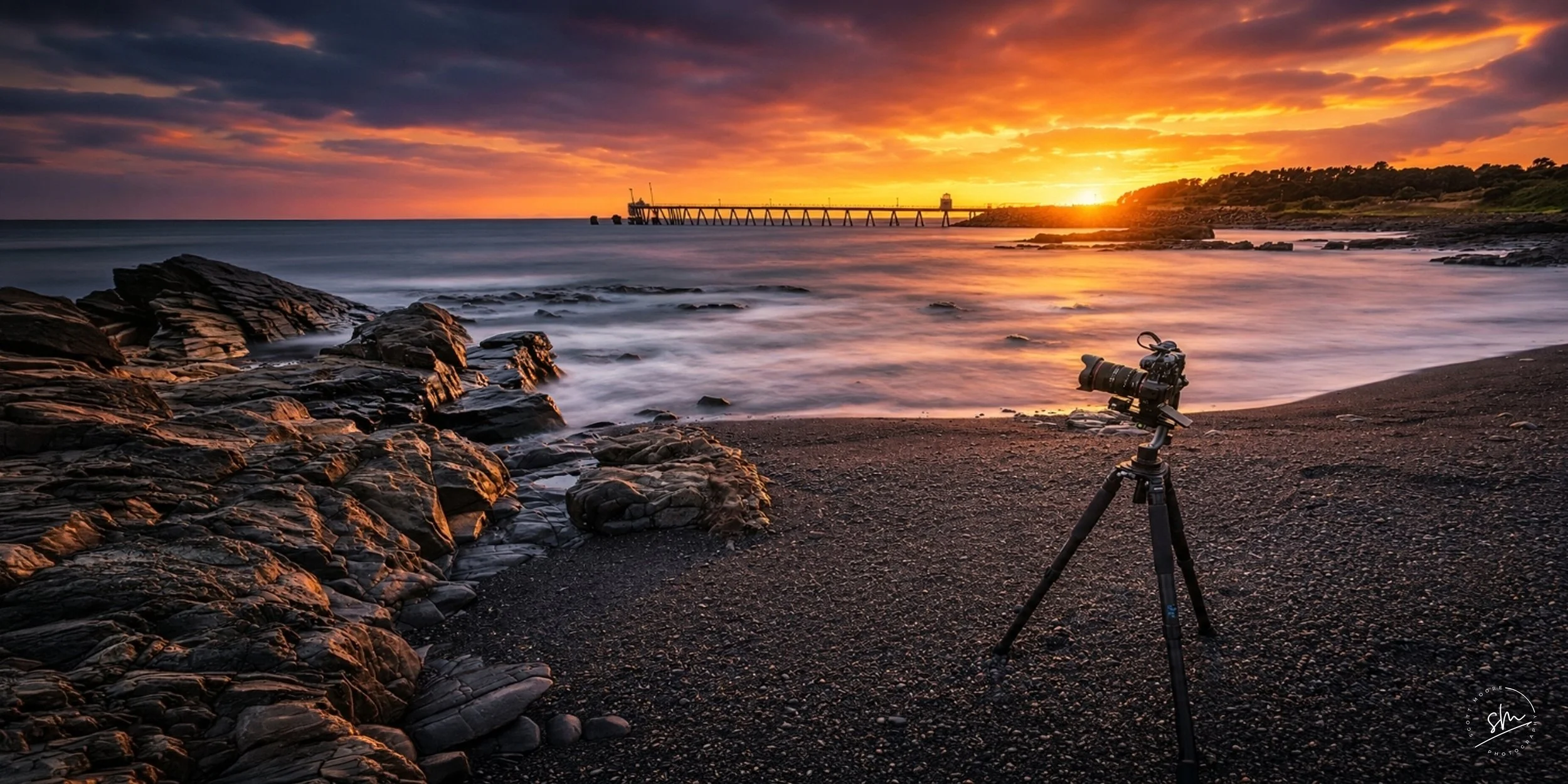 Camera set up on tripod on a rocky beach during sunset with a view of the ocean and a pier extending into the water.