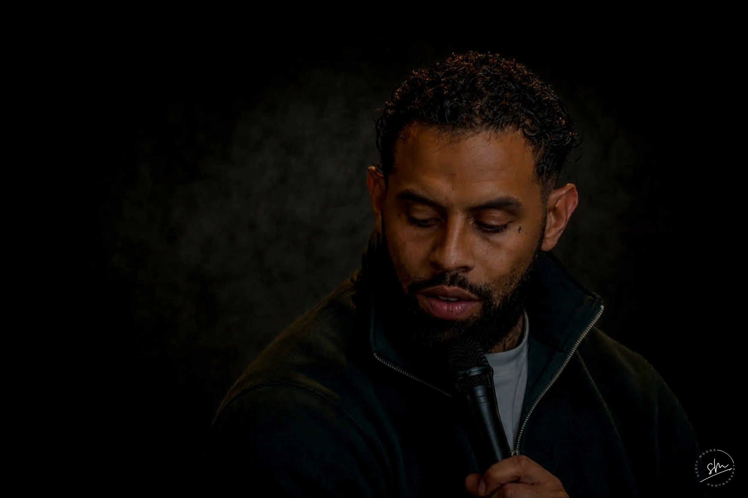 A man with dark, curly hair and a beard looking down, wearing a dark jacket and a light gray shirt, holding an umbrella, against a dark background.