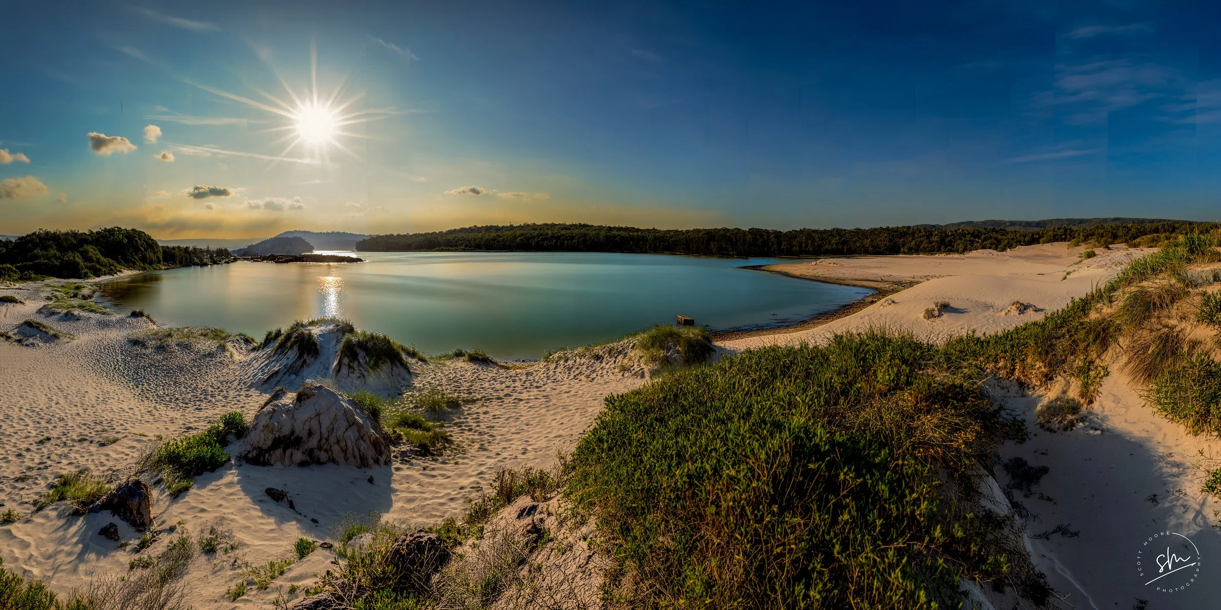 Sunlit river flowing through sandy banks with green vegetation, under a bright sky with scattered clouds and the sun shining.