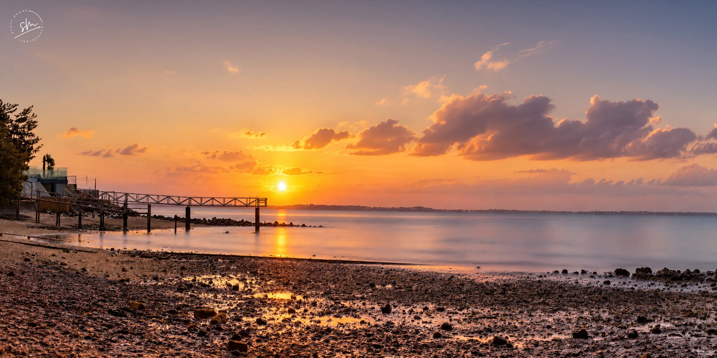 Sunset over a calm body of water with clouds in the sky, a rocky shoreline, and a pier extending into the water on the left side.