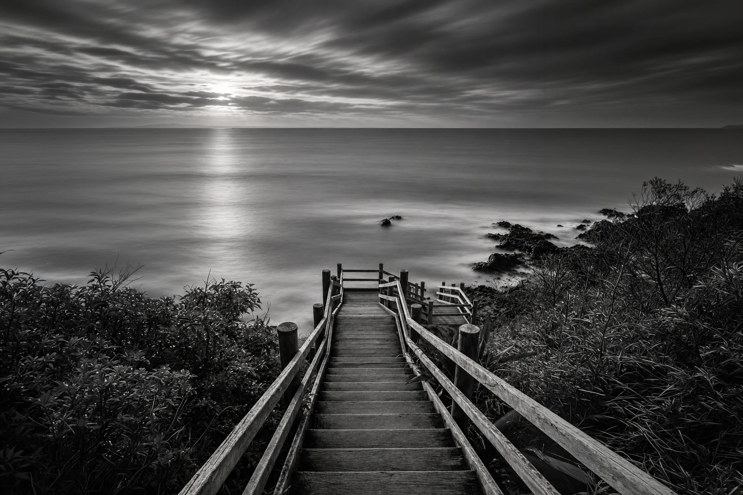 A black and white photo of a wooden staircase leading down to a rocky shoreline with trees, over a calm body of water under a cloudy sky, with the sun partially visible.