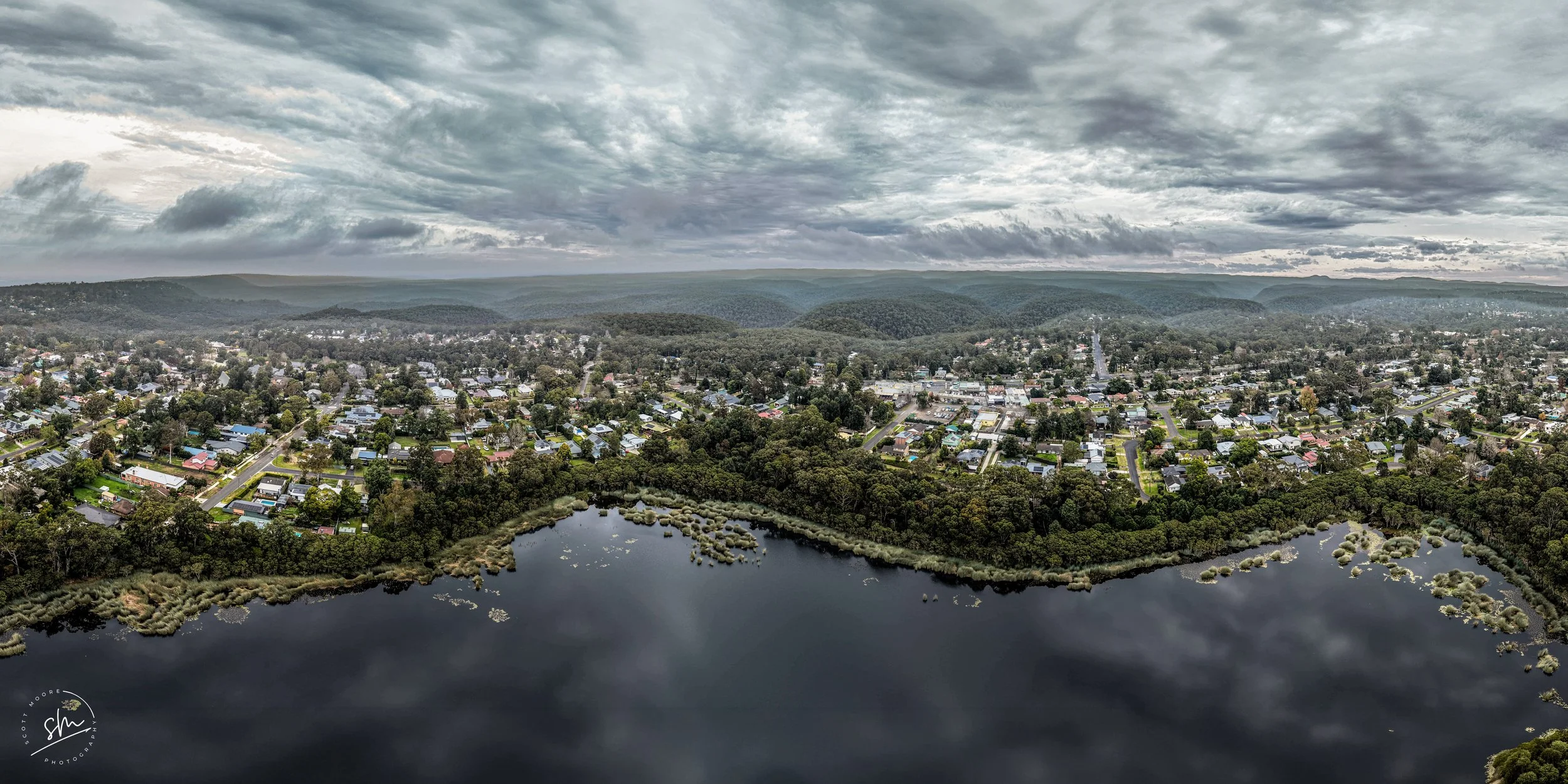 Aerial view of a suburban area with numerous houses, a small lake with vegetation on the edges, and rolling hills in the background under a cloudy sky.