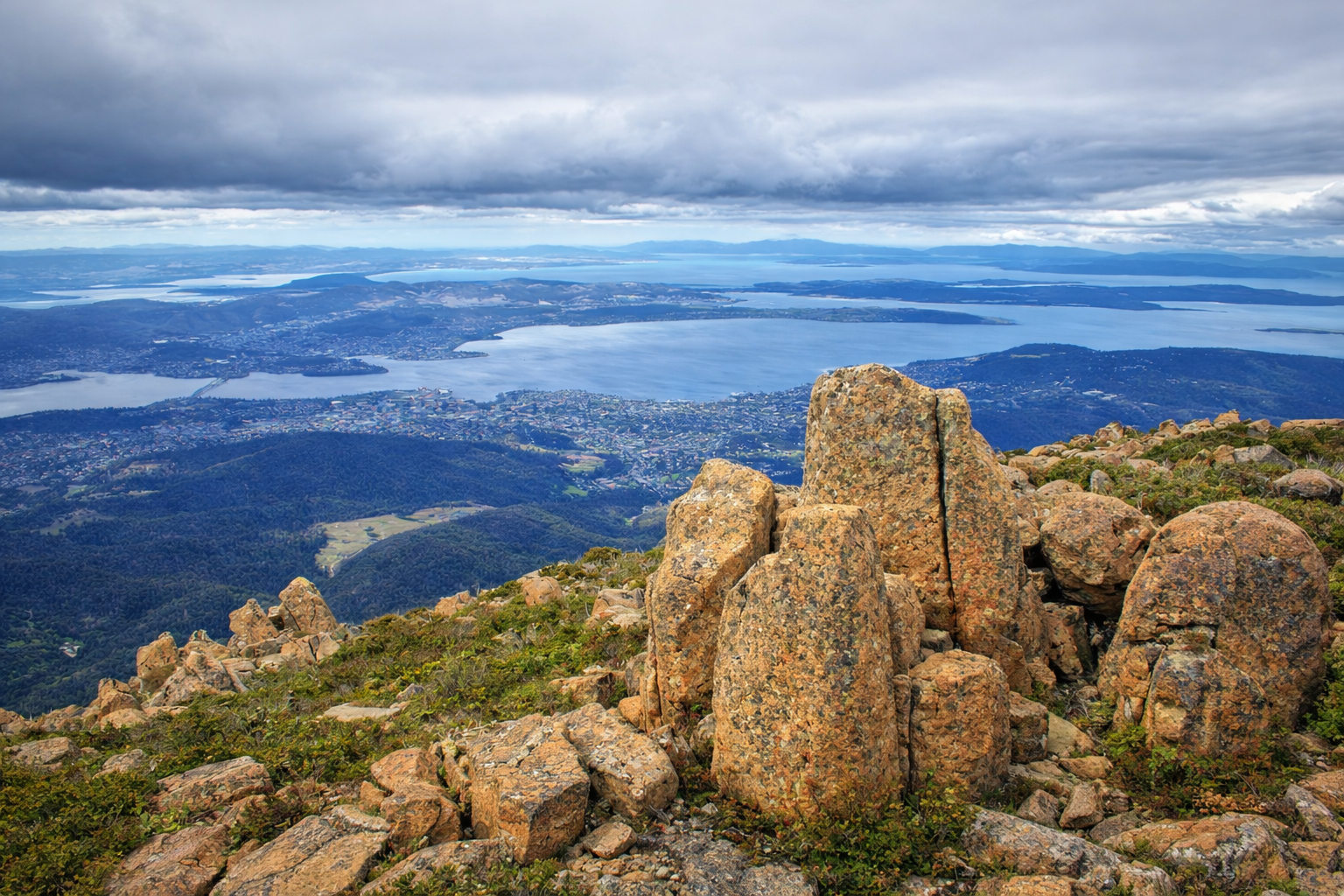 View from a mountain summit overlooking a lake, with large rocks in the foreground, green hills, and a cloudy sky.