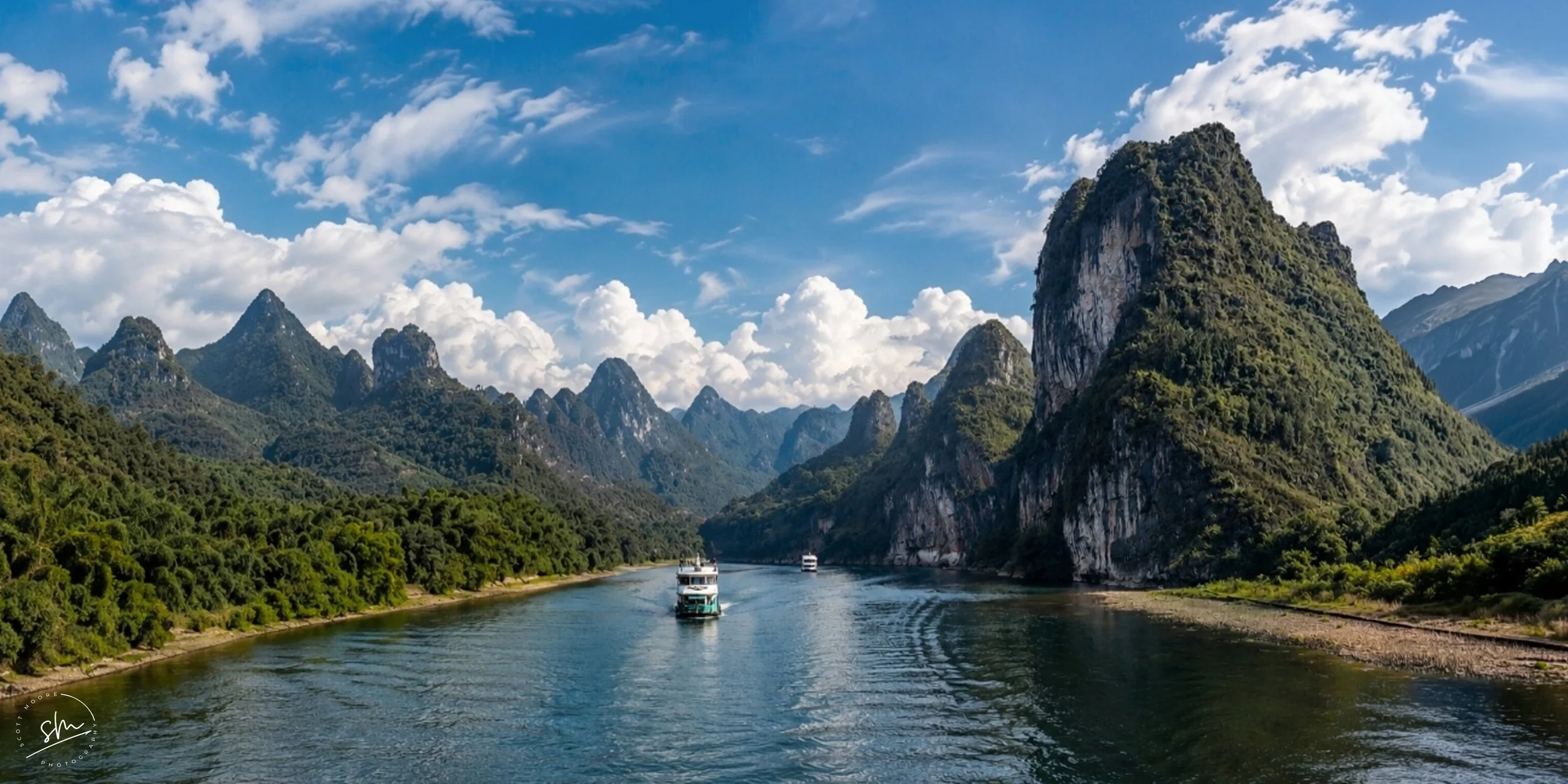 A scenic river surrounded by lush green mountains and tall rocky cliffs under a partly cloudy blue sky with boats sailing on the water.