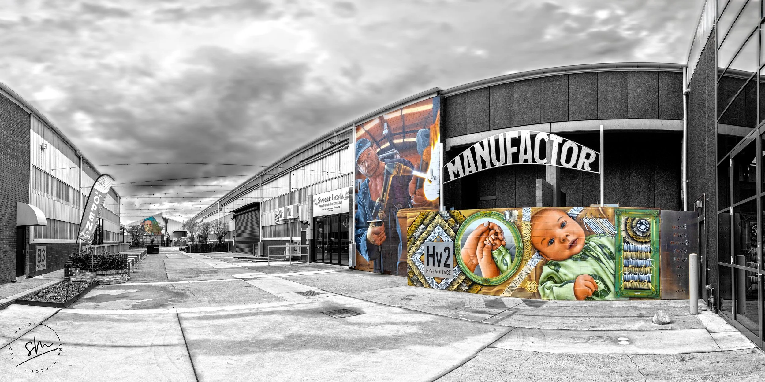 An industrial building with colorful murals, including a construction worker with a hammer, a smiling baby, and various artwork, under a cloudy sky.