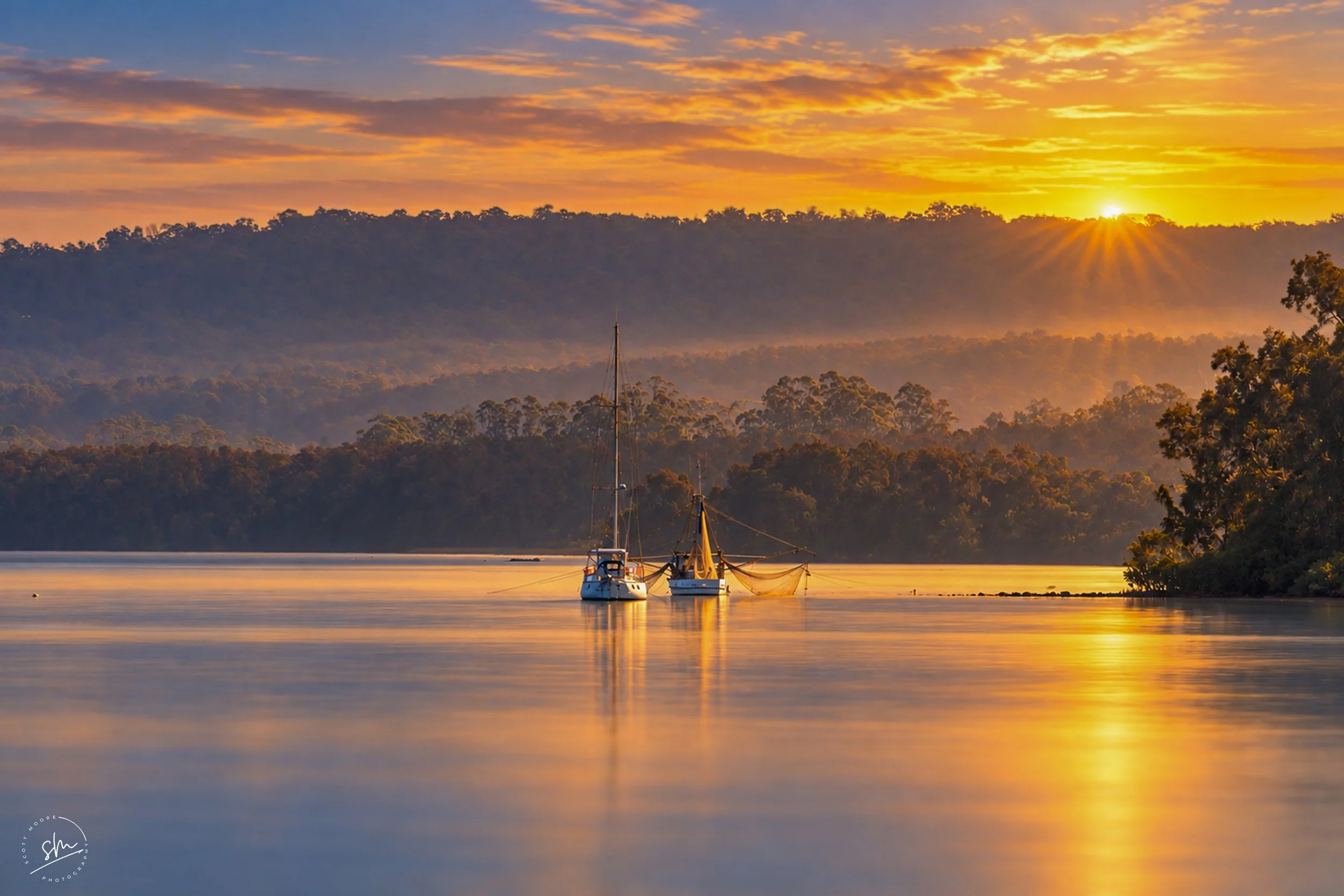 A serene lakeside scene at sunset with two boats anchored on calm water, surrounded by forested hills, and a sky illuminated with orange and yellow clouds.