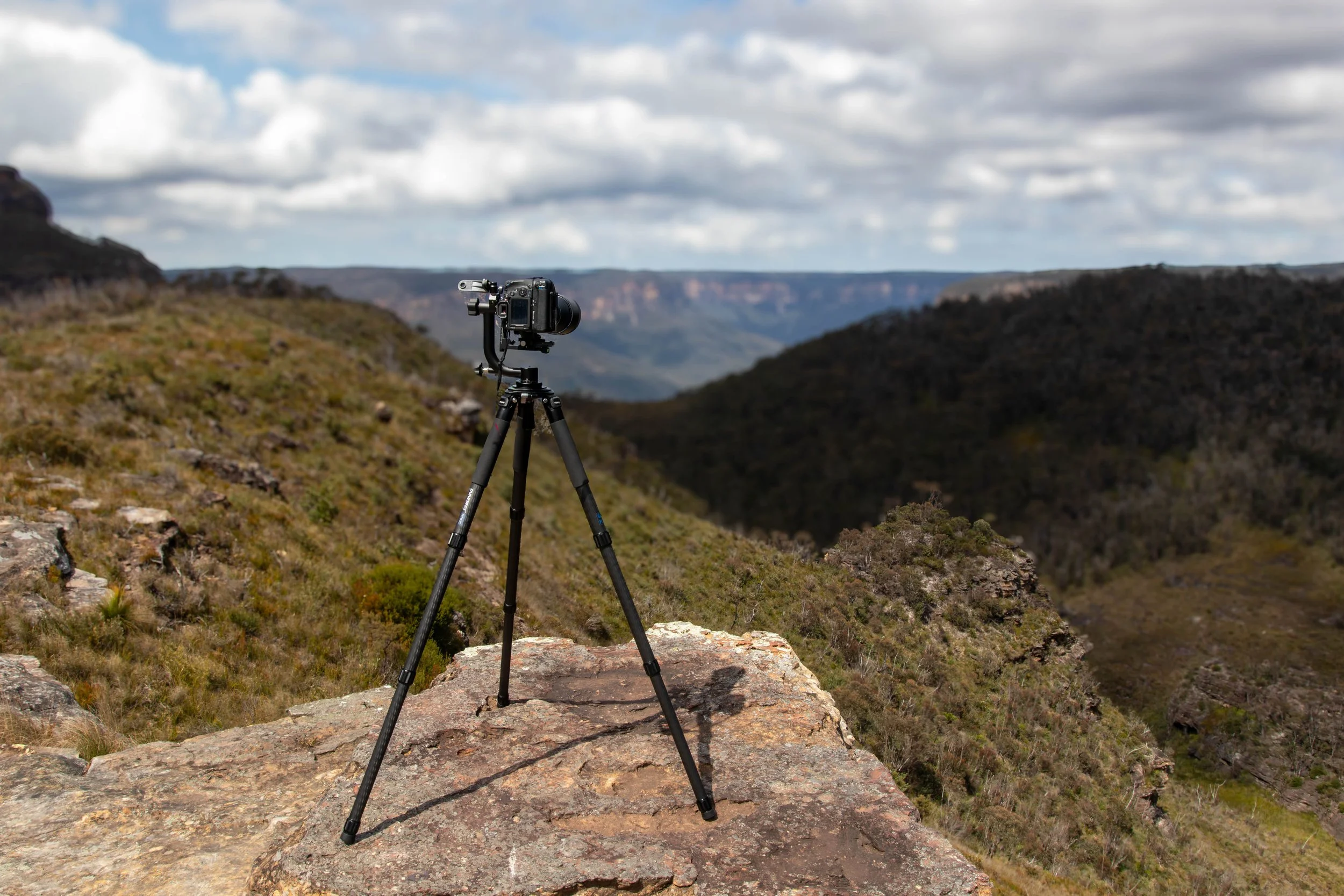 A digital camera mounted on a tripod positioned on a rocky ledge overlooking a canyon landscape with rolling hills and a cloudy sky.