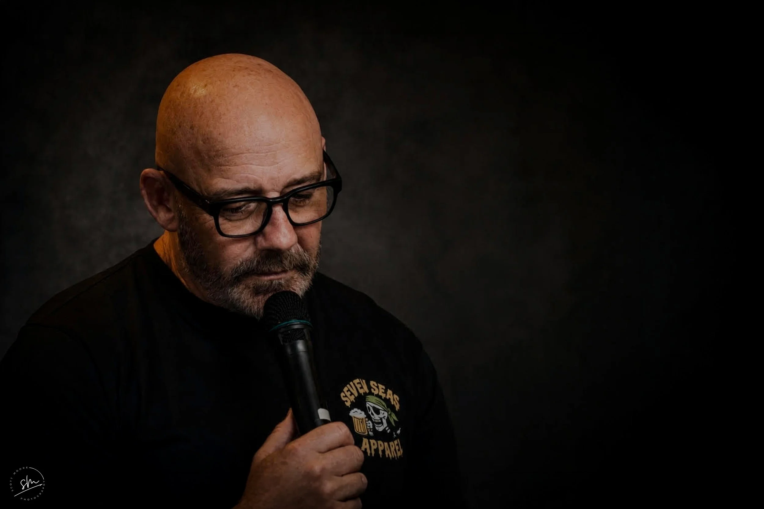 A bald, bearded man wearing glasses, holding a microphone, and wearing a black t-shirt with a graphics logo, against a black background.