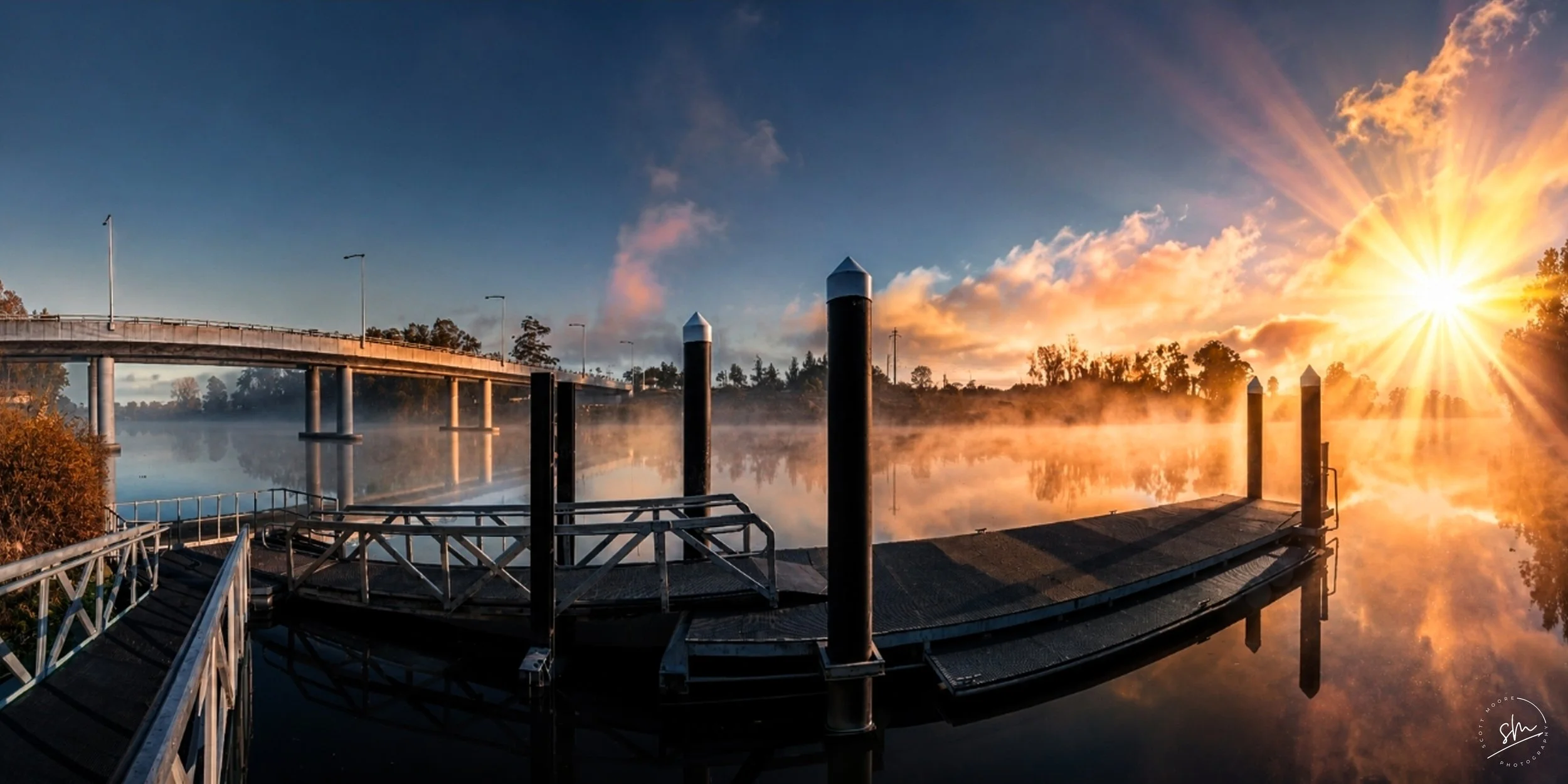 Sunrise over a calm river with a bridge and dock, mist rising from the water, trees in the background, and a clear sky with a few clouds.