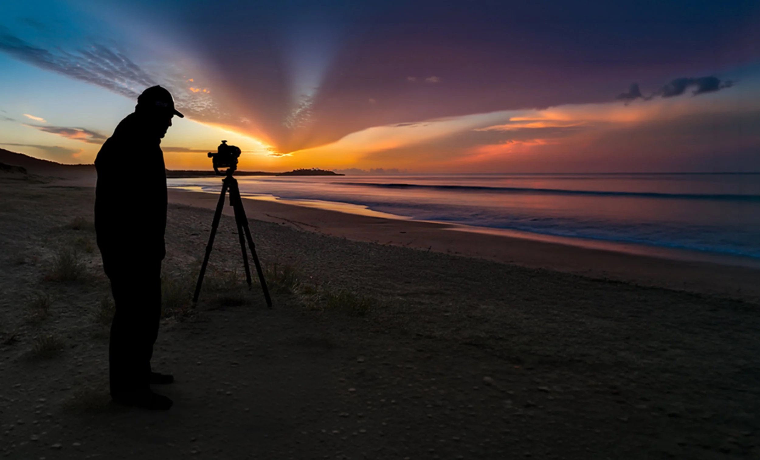 Silhouette of a person with a camera on a tripod capturing a colorful sunset or sunrise over the ocean on a beach.