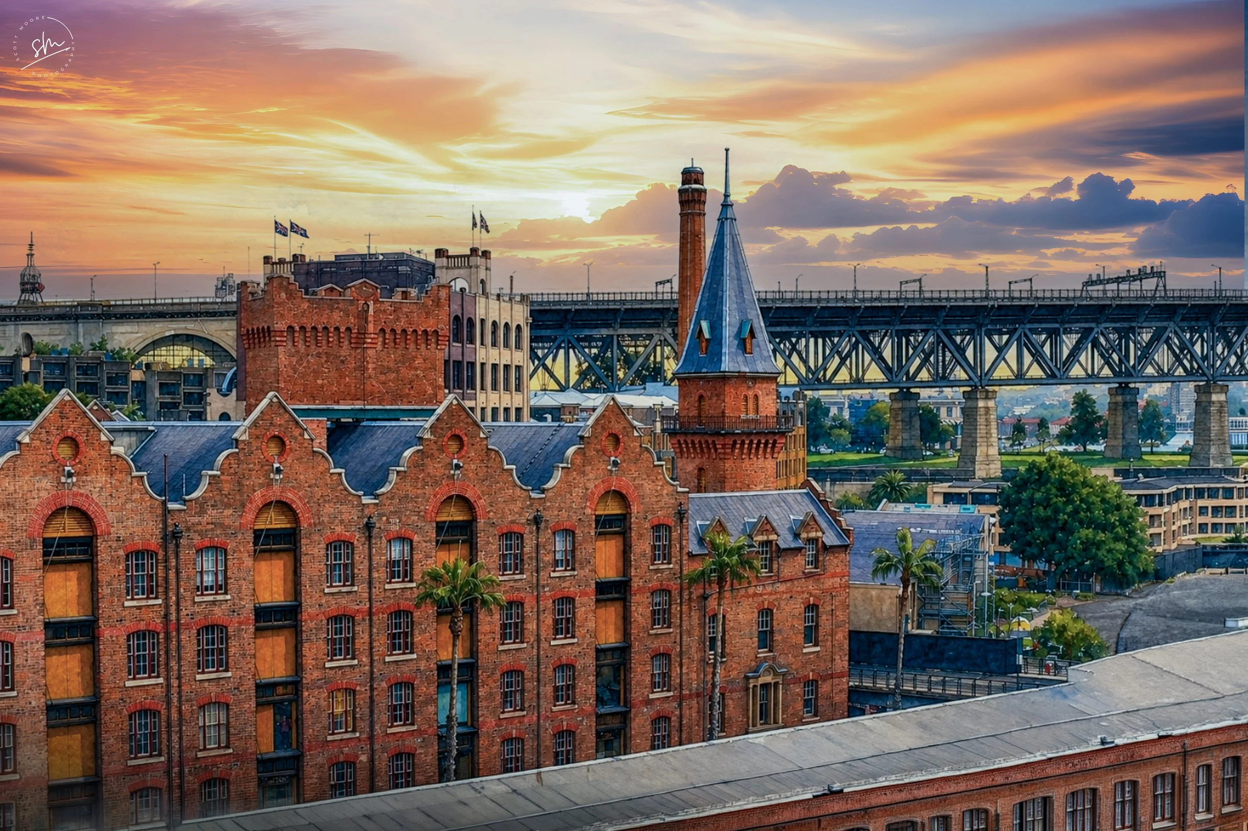 Cityscape at sunset with historic red brick buildings, a church with a tall steeple, a bridge in the background, and a colorful sky with clouds.