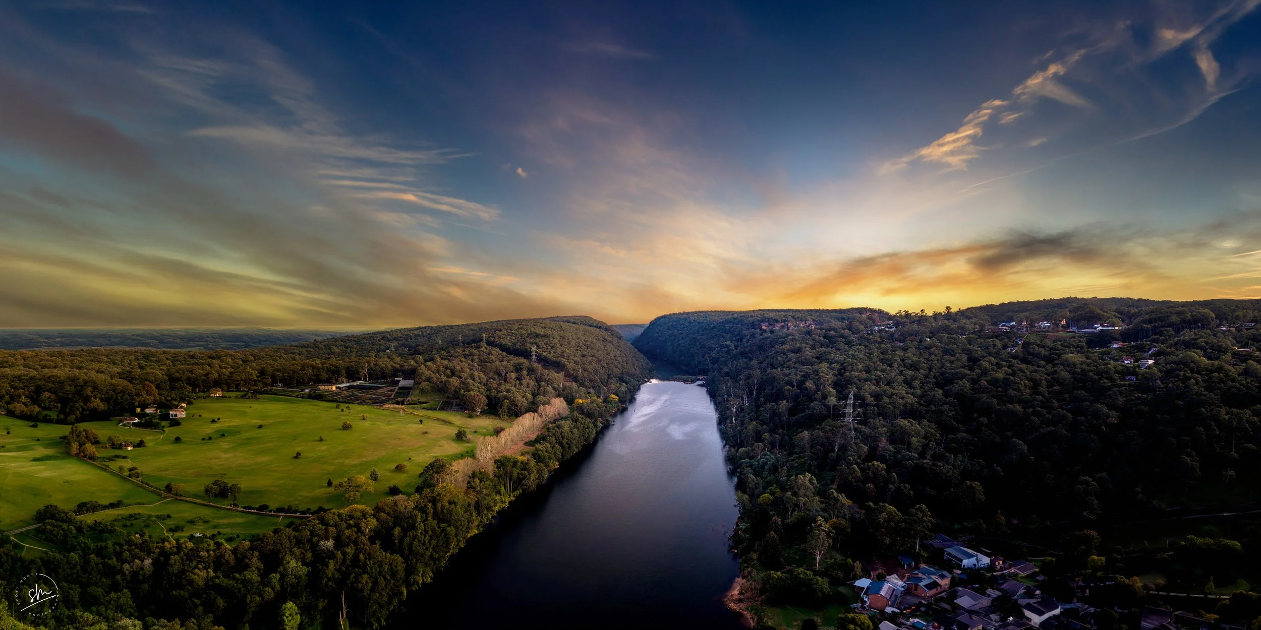 View of a river flowing between forested hills during sunset with a partly cloudy sky.