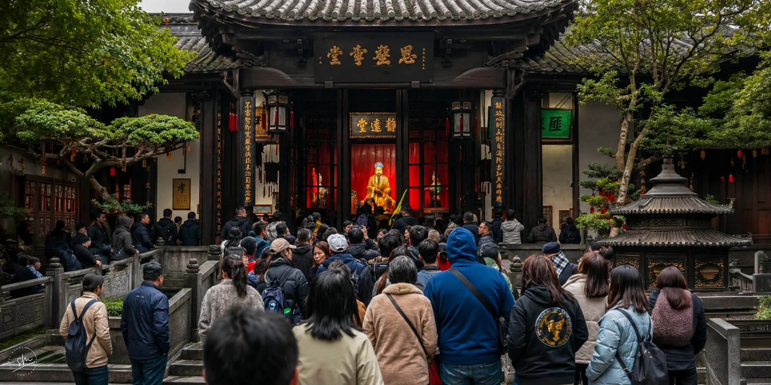 Crowd of people gathered in front of a traditional Chinese temple with a golden deity statue inside, surrounded by trees and wooden architecture.