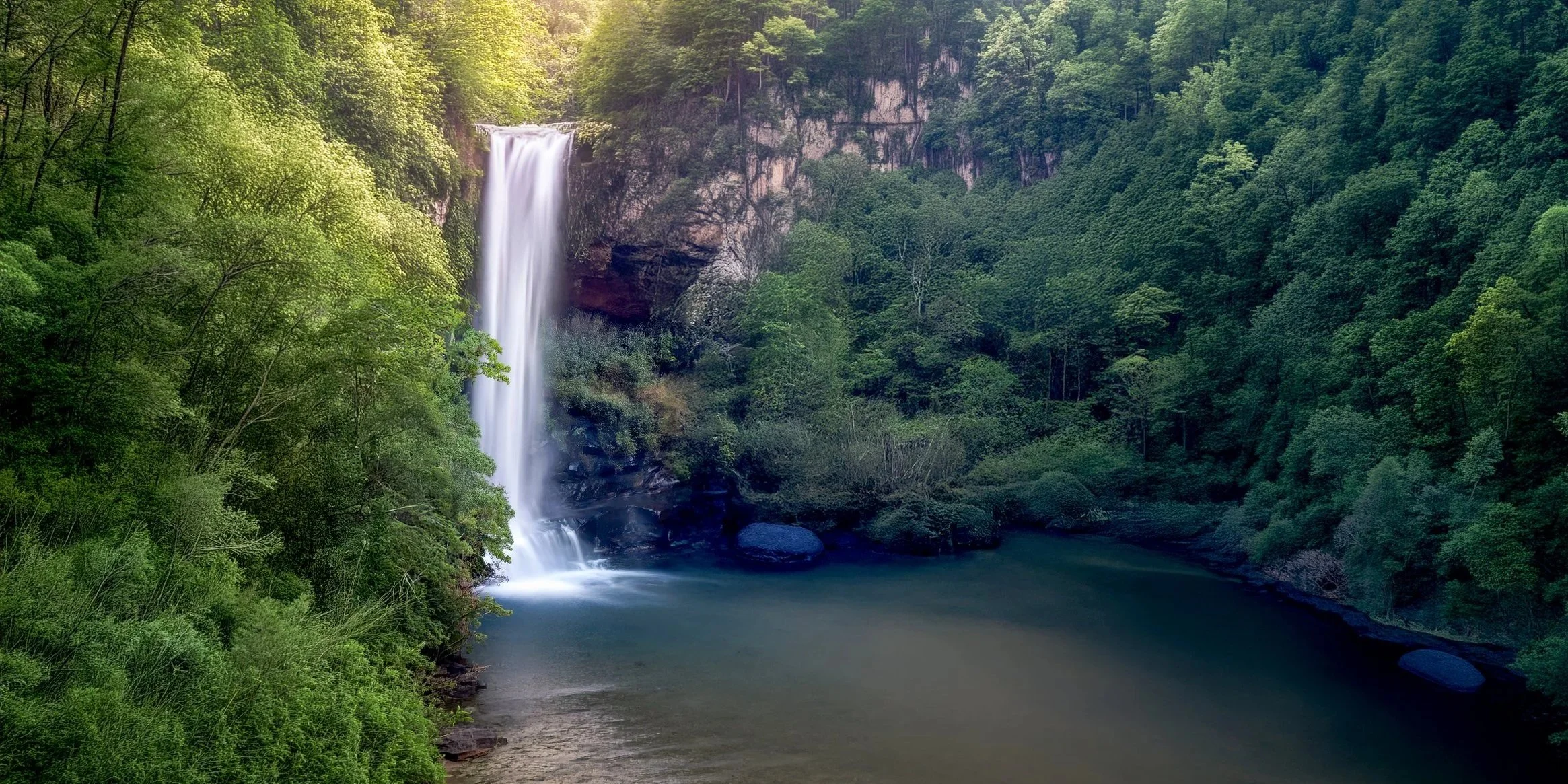 A waterfall flowing down a cliff into a river surrounded by dense green trees and foliage.