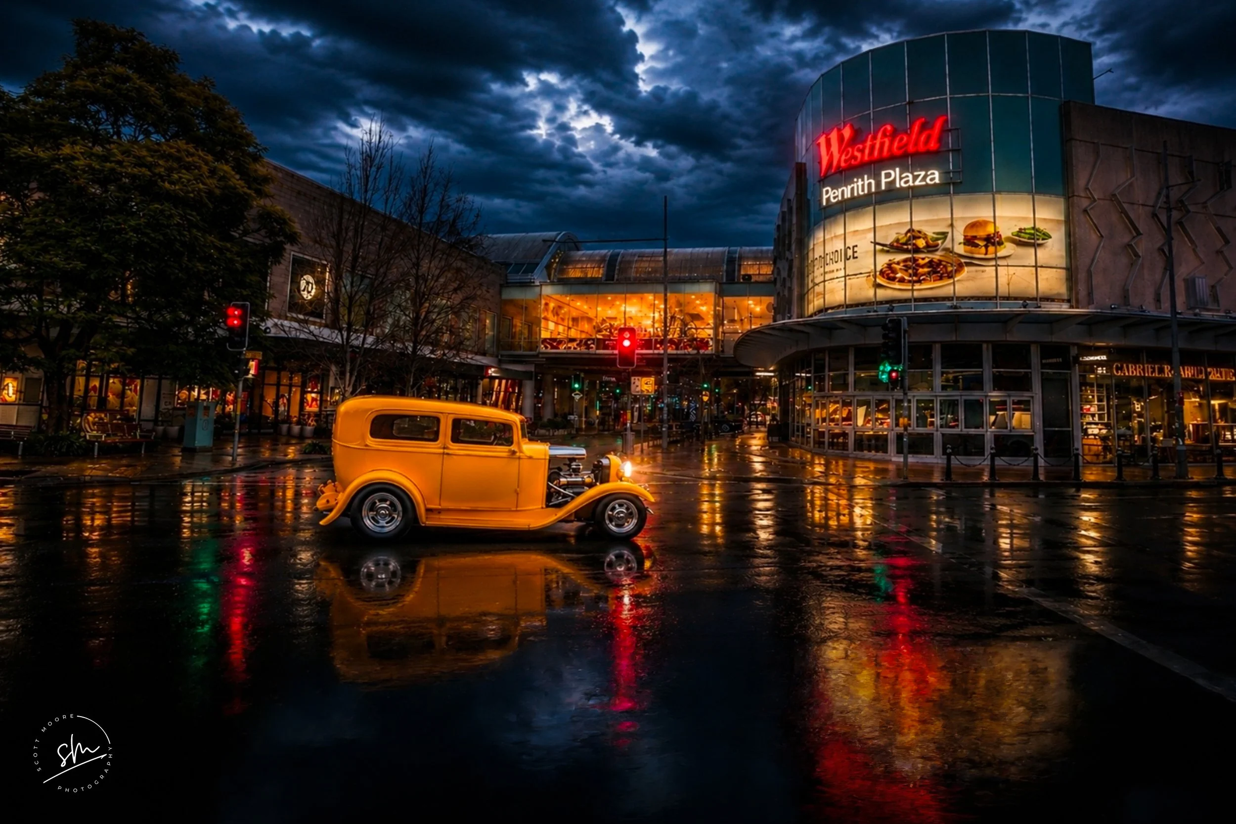 A vintage yellow car parked in front of Westfield Penrith Plaza shopping mall at night with dark stormy clouds overhead. The mall's illuminated sign and interior lights reflect off the wet pavement.