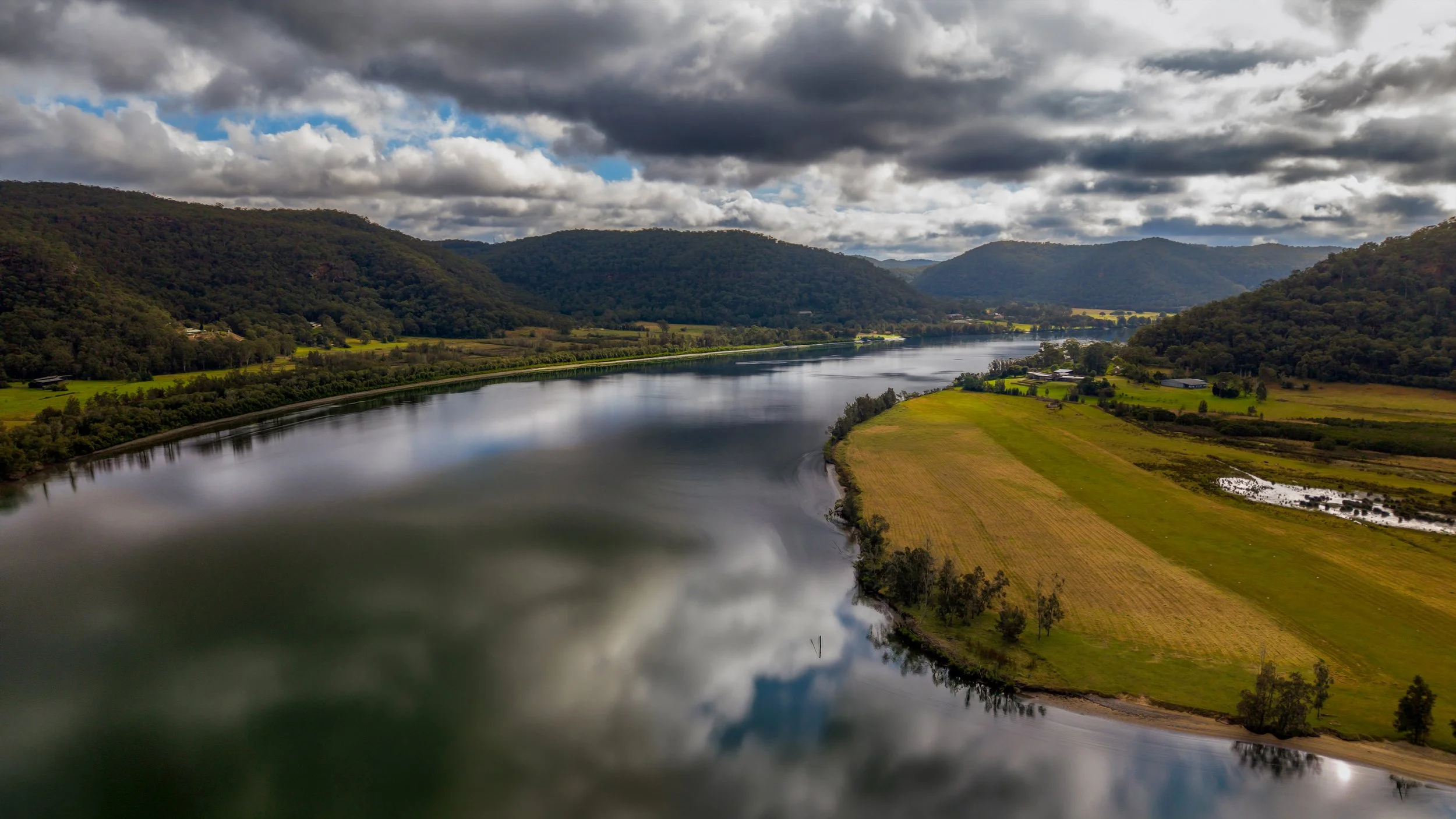A large river flows through a valley surrounded by green hills and mountains under a cloudy sky.