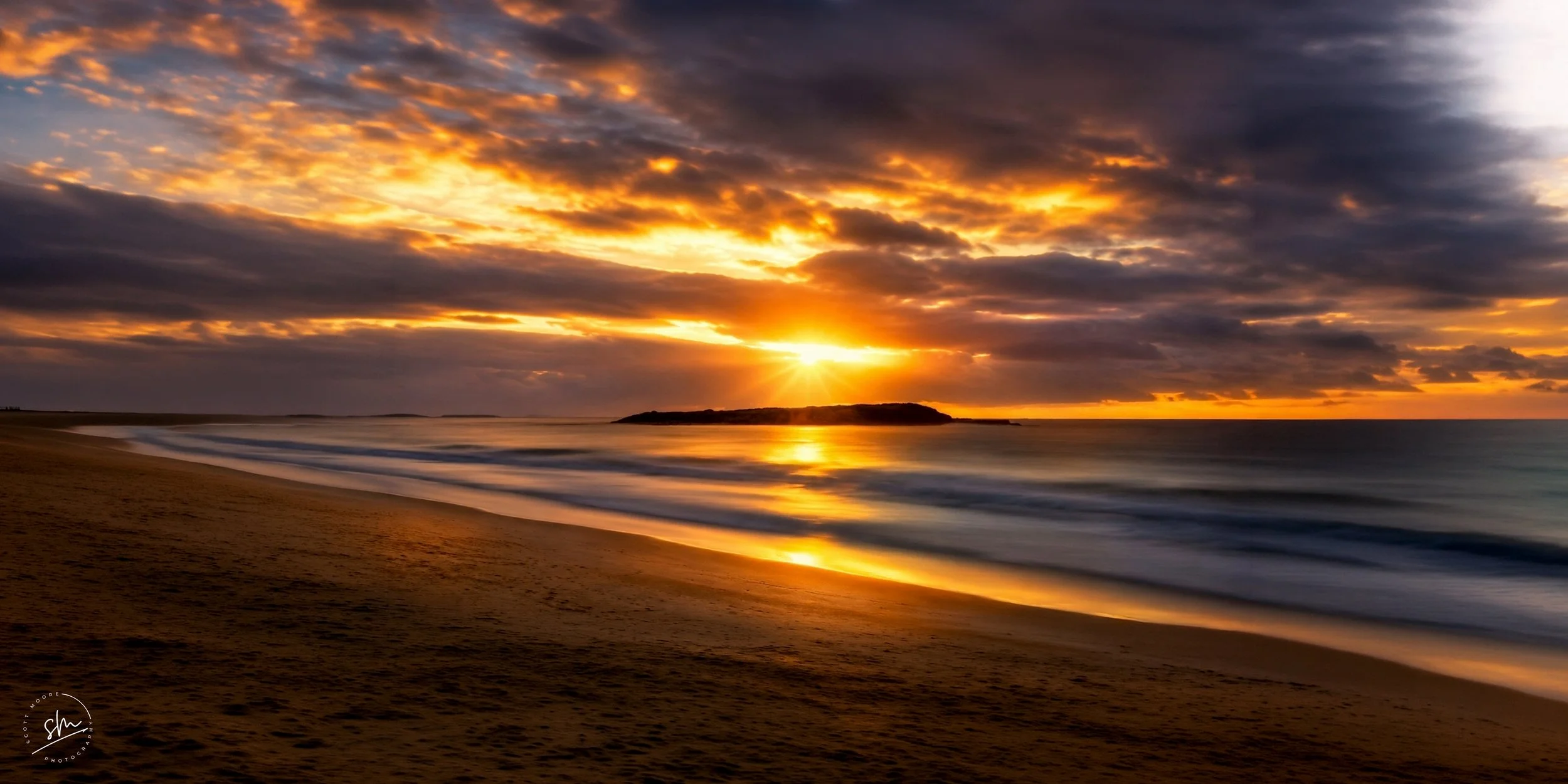 Sunset over the ocean with a sandy beach in the foreground and a small island on the horizon, clouds reflecting the warm colors of the setting sun.