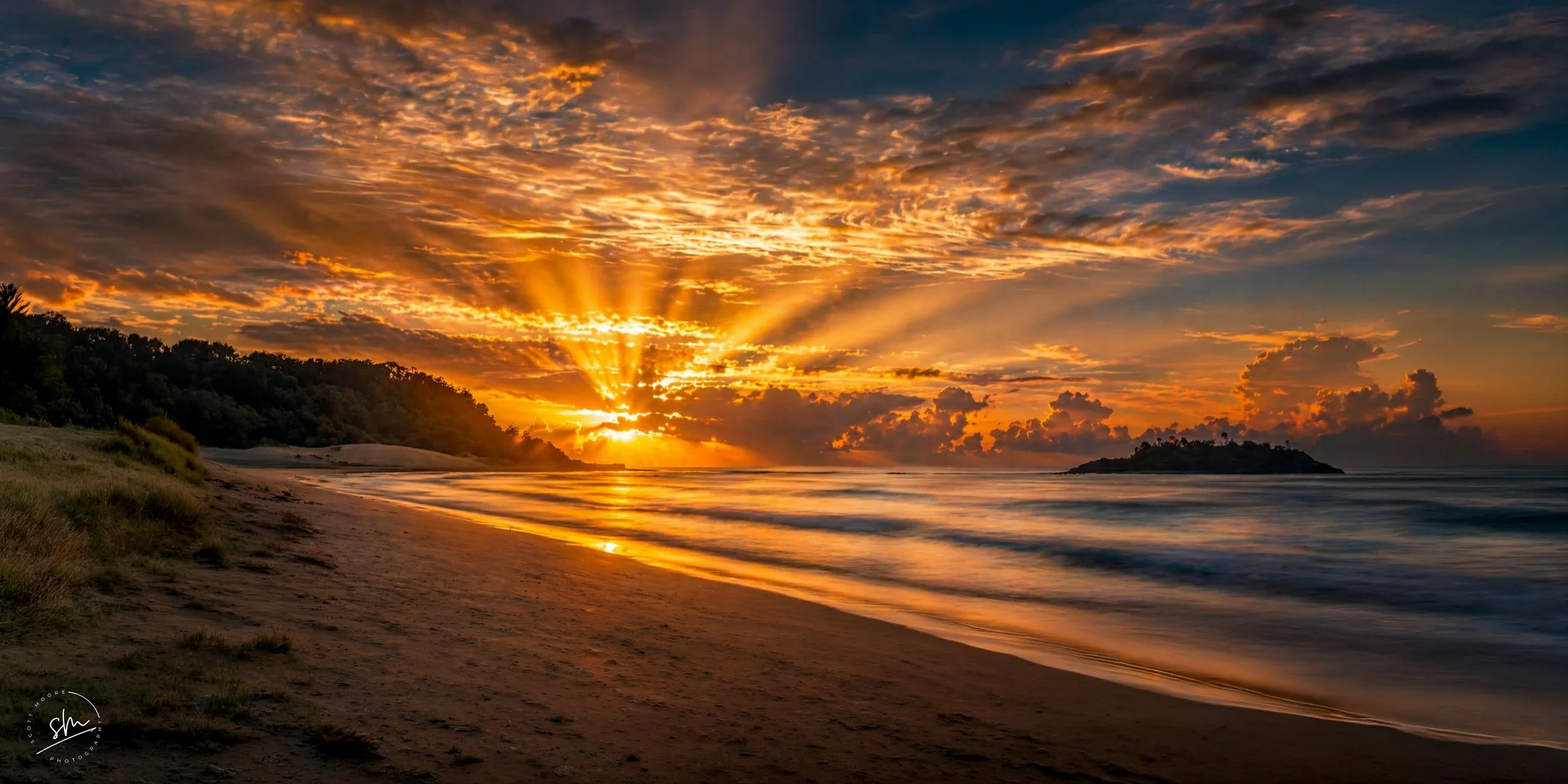Sunset over a beach with cloudy sky, water, and small island in the distance.
