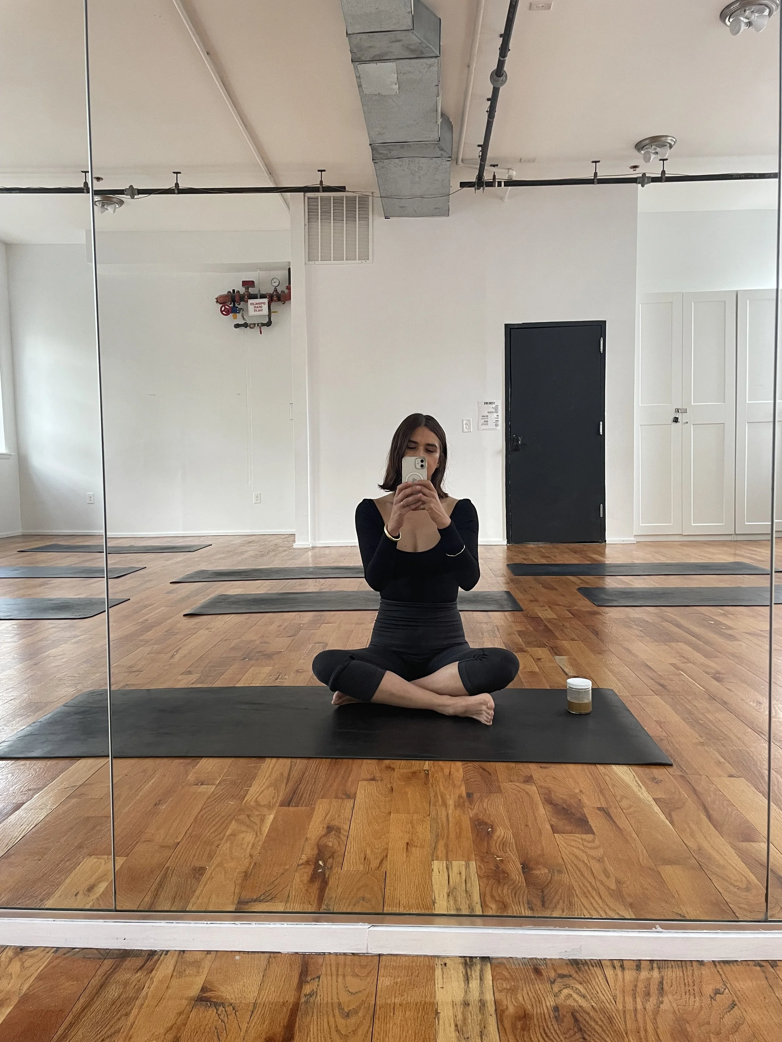A woman sitting cross-legged on a yoga mat in a studio, taking a mirror selfie with her phone. The studio has wooden flooring, a mirrored wall, and minimalist decor, including yoga mats and a small jar container in front of her.