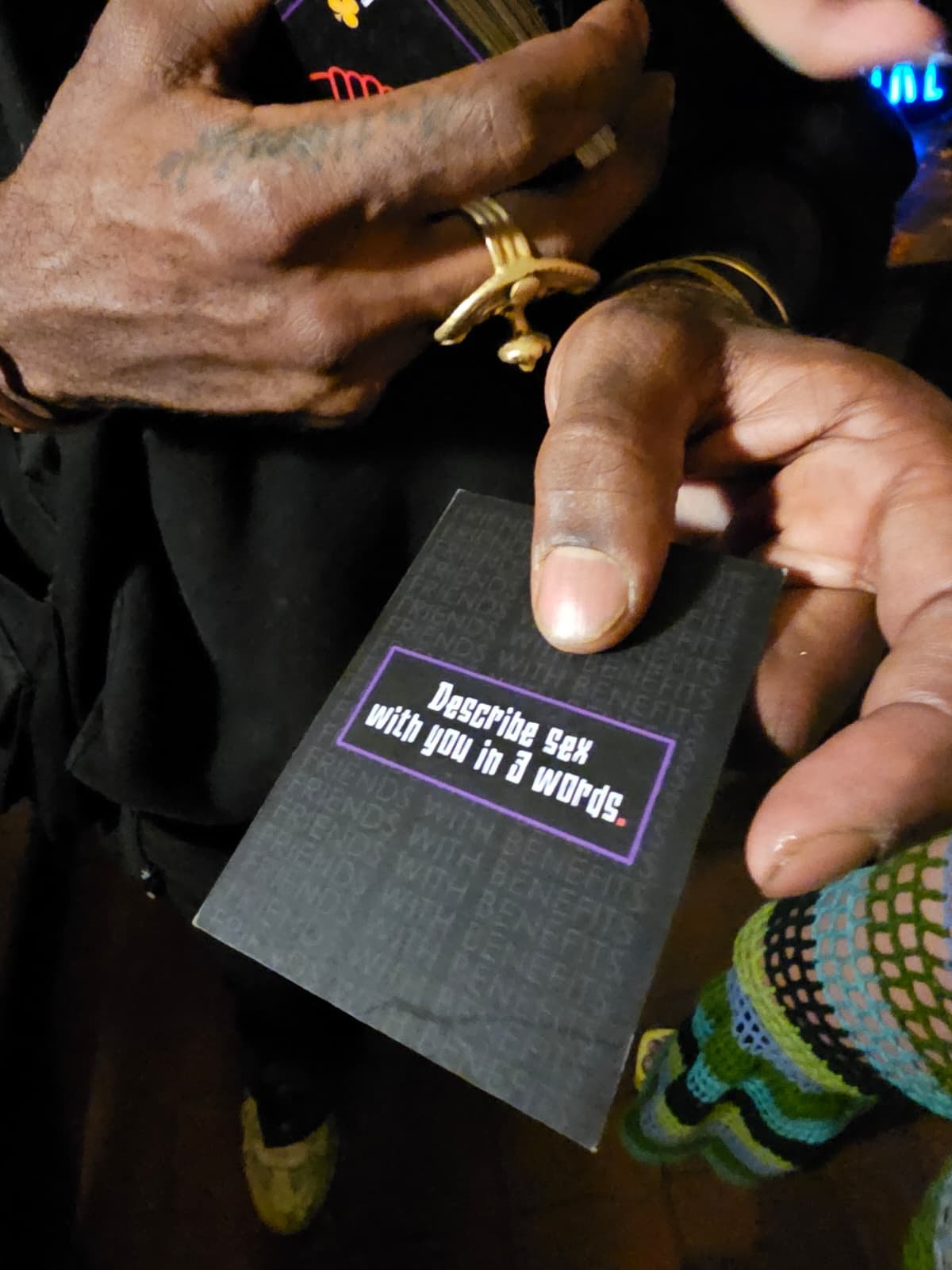A person with tattoos on their hand holds a black card that says, 'Describe sex with you in three words.'