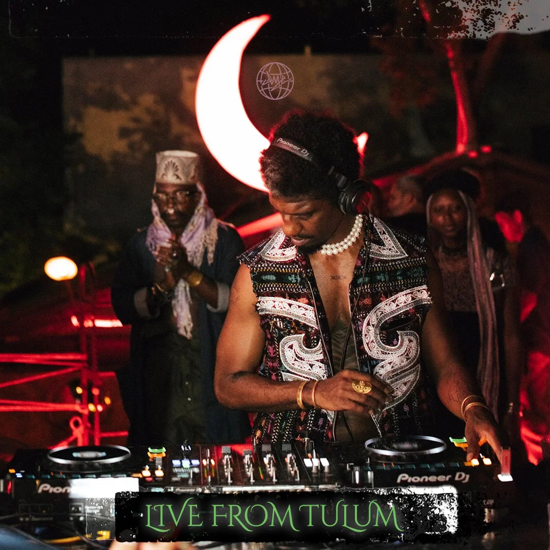 A DJ wearing a beaded necklace and a patterned vest operates turntables highlighted with colorful lights in a dim club with red and black lighting. Behind the DJ is a cosmic moon-shaped neon light, with people dancing in the background, and a sign reading 'Live from Tulum' at the bottom.