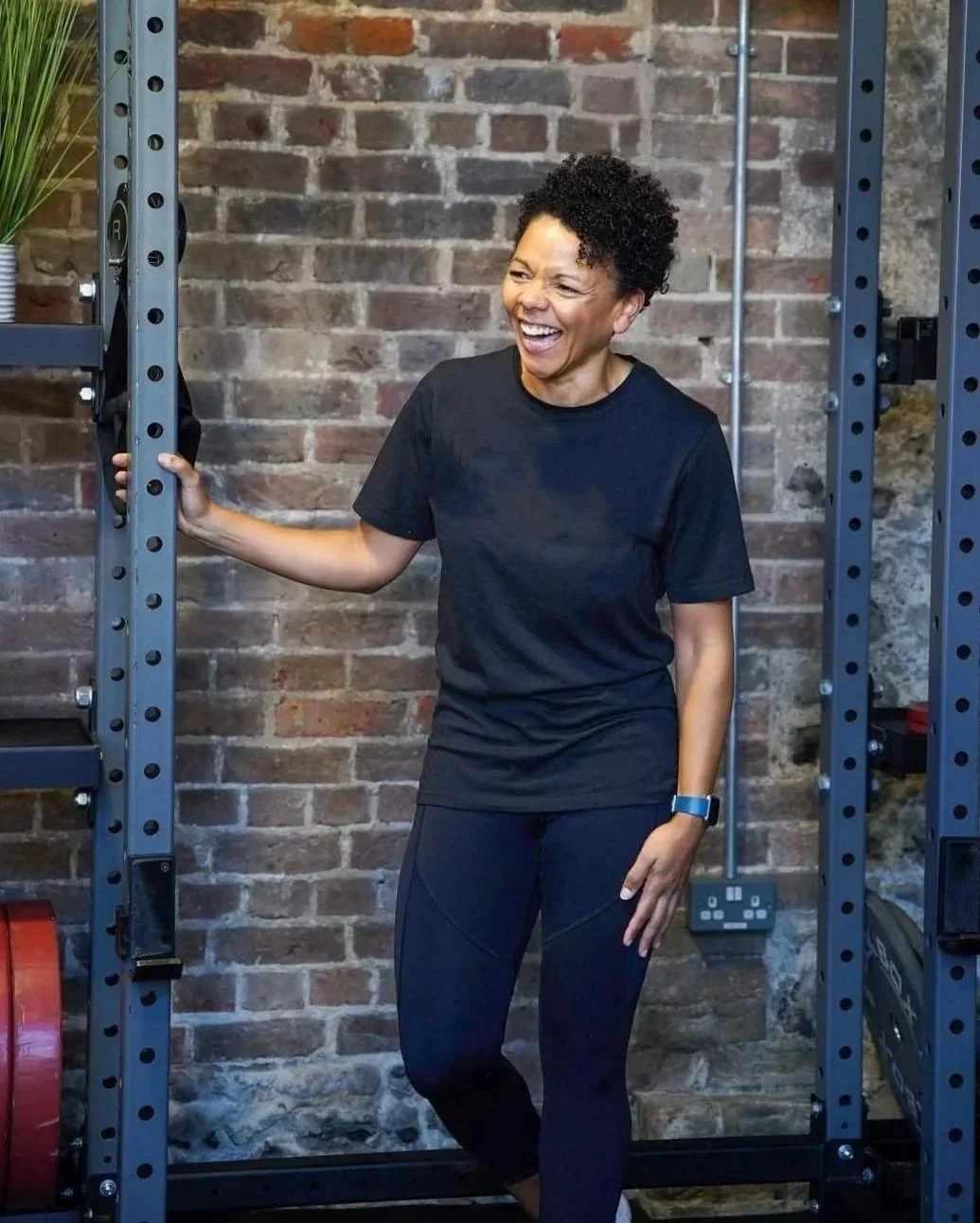 A photo of a brown woman laughing and smiling she is leaning against a gym rack with a brick wall in the background