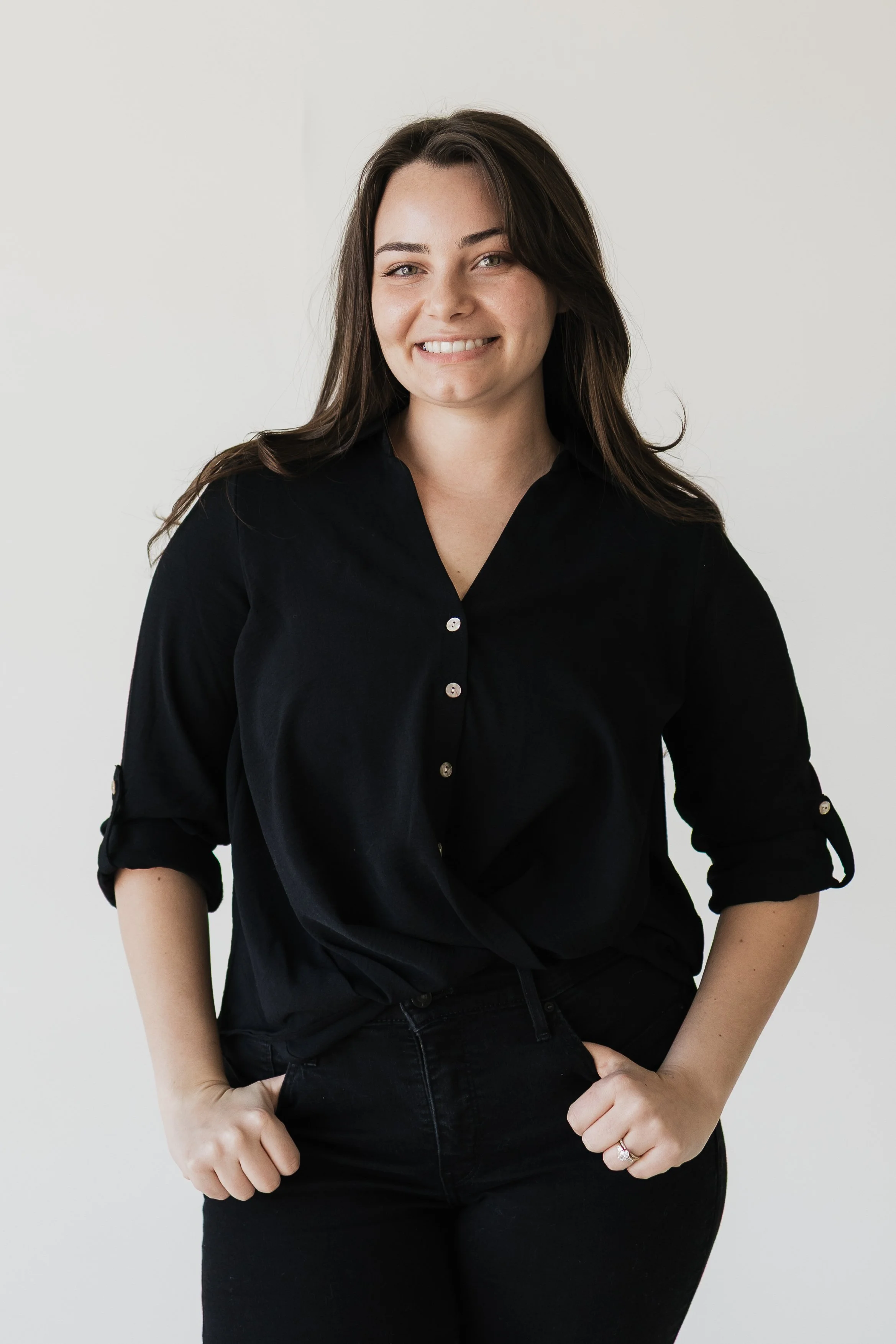 A woman with long brown hair smiling and wearing a black button-up shirt with rolled-up sleeves against a plain light background.