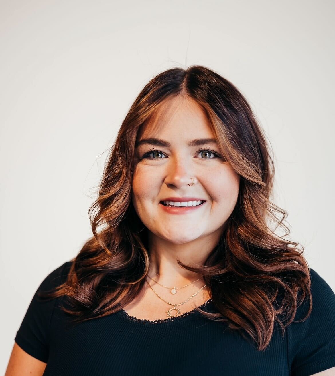 A woman with long, wavy brown hair, blue eyes, and light makeup, smiling in front of a plain light-colored wall.