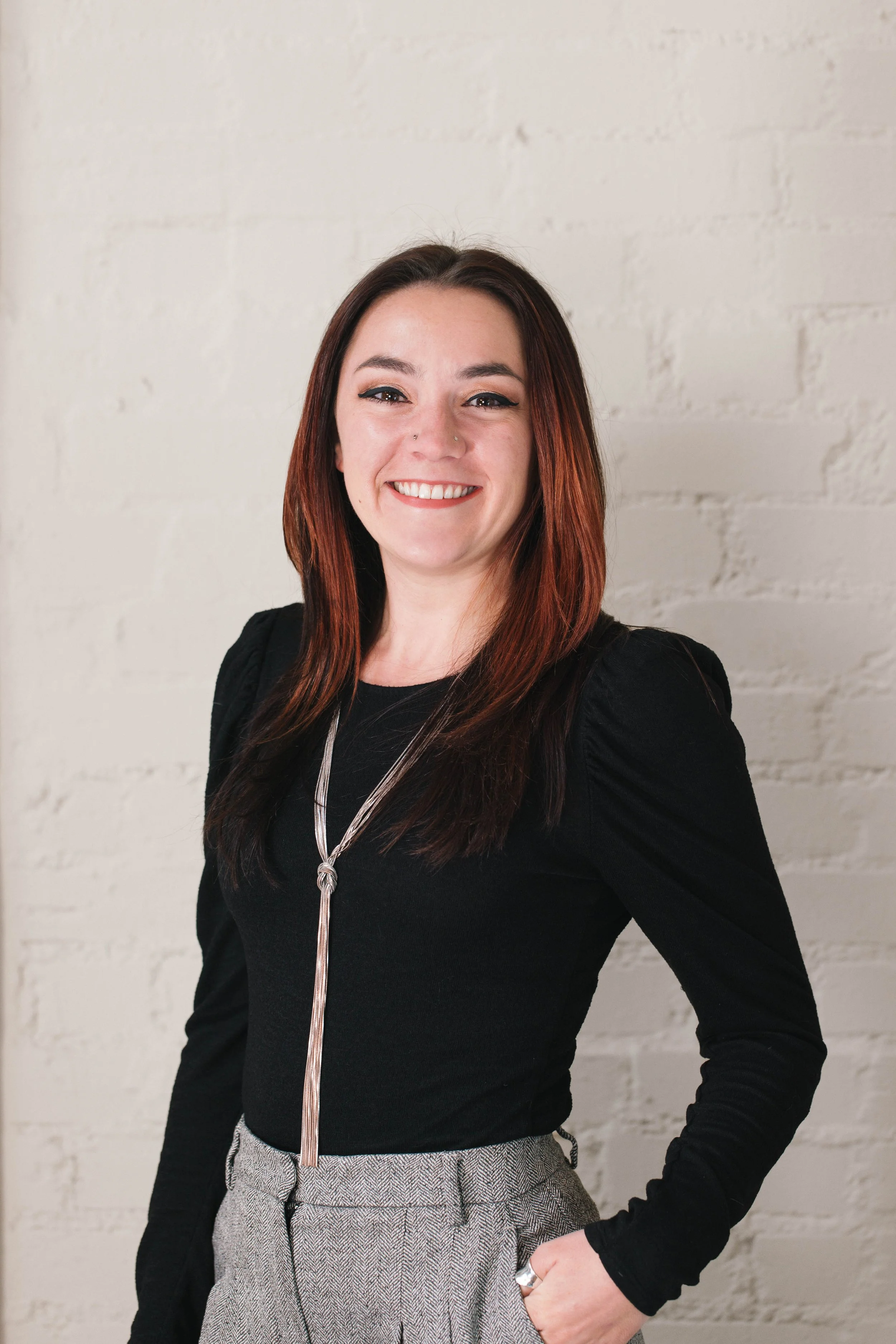 A smiling woman with long, reddish-brown hair wearing a black top and gray plaid pants standing against a white brick wall.
