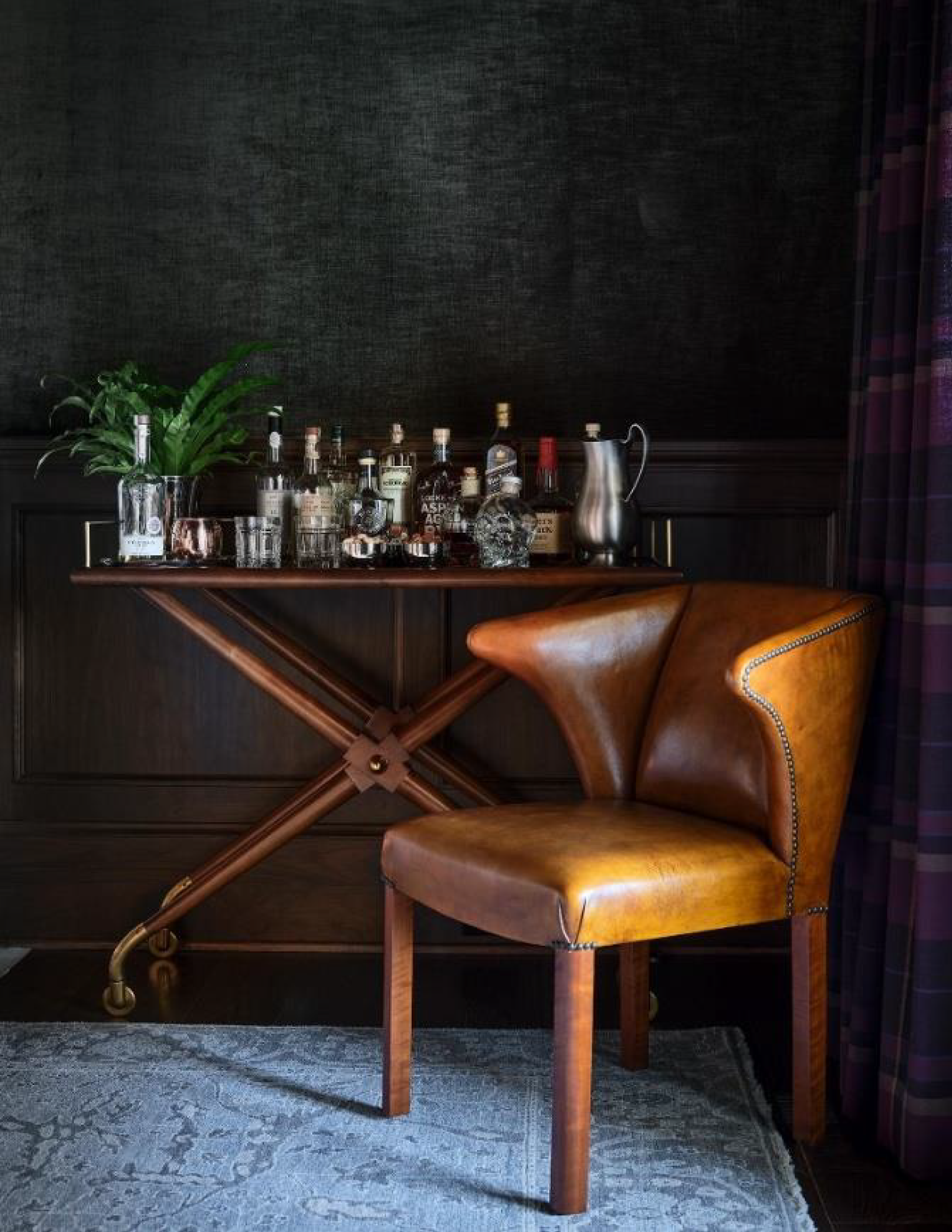 A vintage-style bar cart with liquor bottles and glassware, a potted plant, a skull decoration, and a silver milk pitcher, next to a brown leather chair, all set against a dark textured wall with purple curtains.