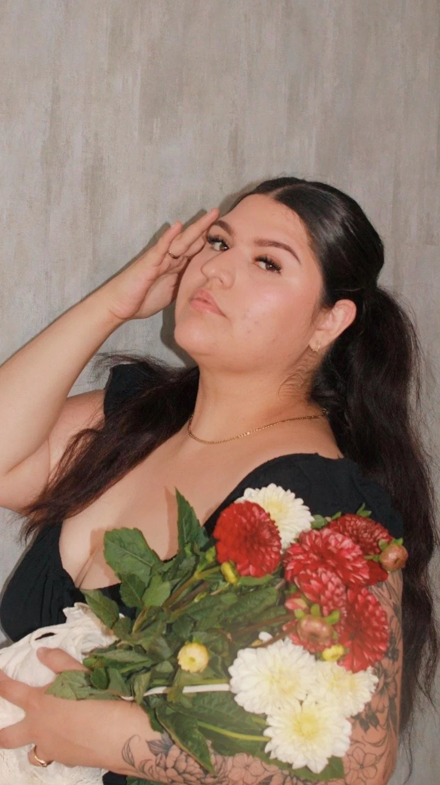 A woman with dark brown hair holding a bouquet of red and white flowers, wearing a black top, standing against a plain background.