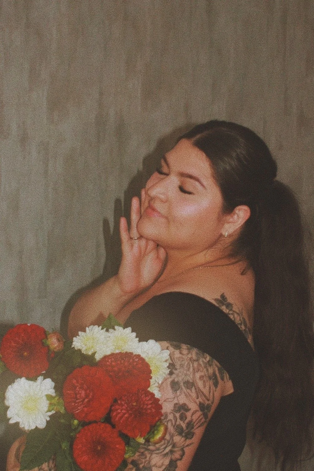 A woman with long dark hair, tattoos on her arm, and earrings, posing with closed eyes and a gentle smile, holding a bouquet of red and white flowers.