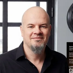 Headshot of a bald man with a goatee, dressed in a black shirt, standing indoors in front of windows.