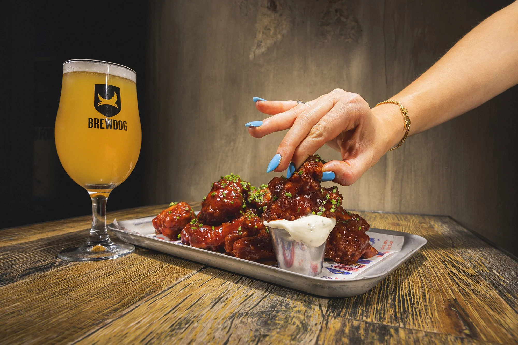 Person with blue painted nails reaching for sauced chicken wings on metal tray with side of ranch dressing, and a glass of beer labeled 'BREWDOG' on wooden table.