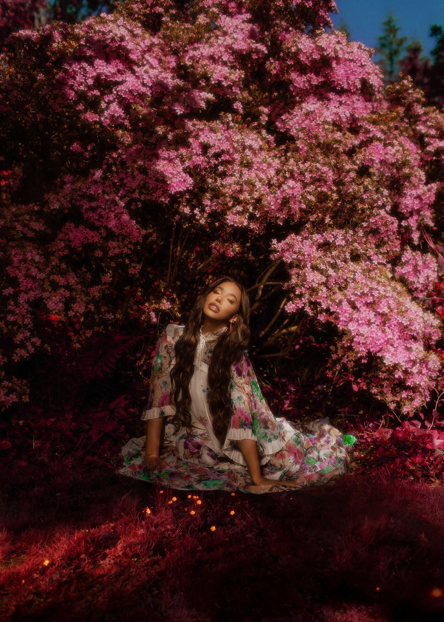 A woman with long wavy hair wearing a floral dress sitting on the ground in front of a large pink flowering shrub at dusk or nighttime.