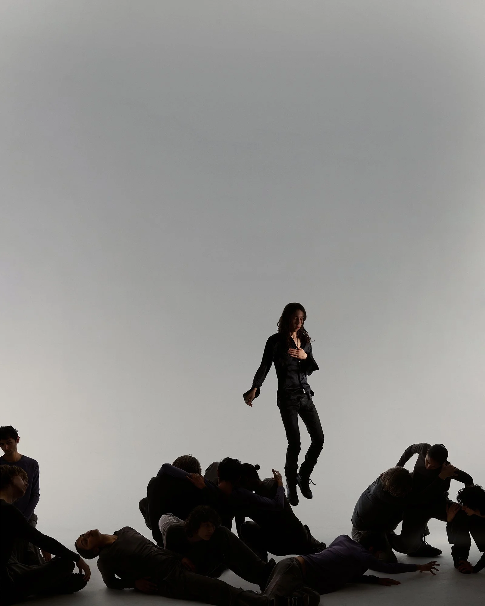 A group of people, with one woman in a black outfit jumping in mid-air above them, set against a plain gray background.