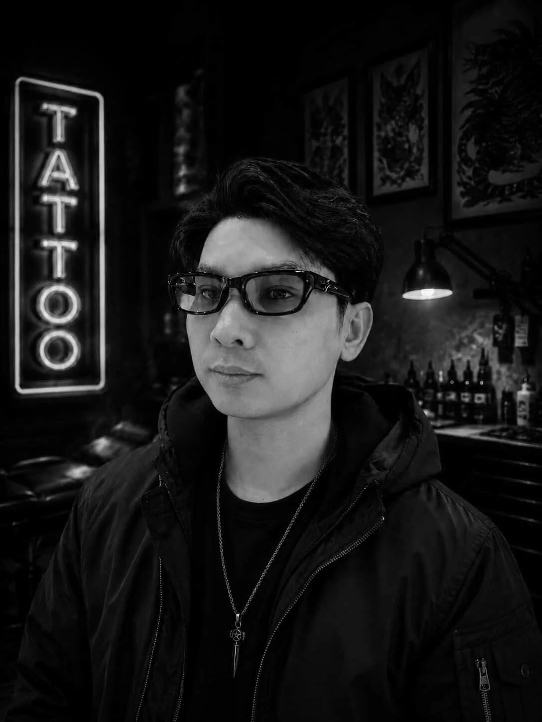 Black and white photo of a young man with dark hair and glasses, wearing a black jacket and necklace, sitting in a tattoo parlor with a sign that reads 'TATTOO' in the background.