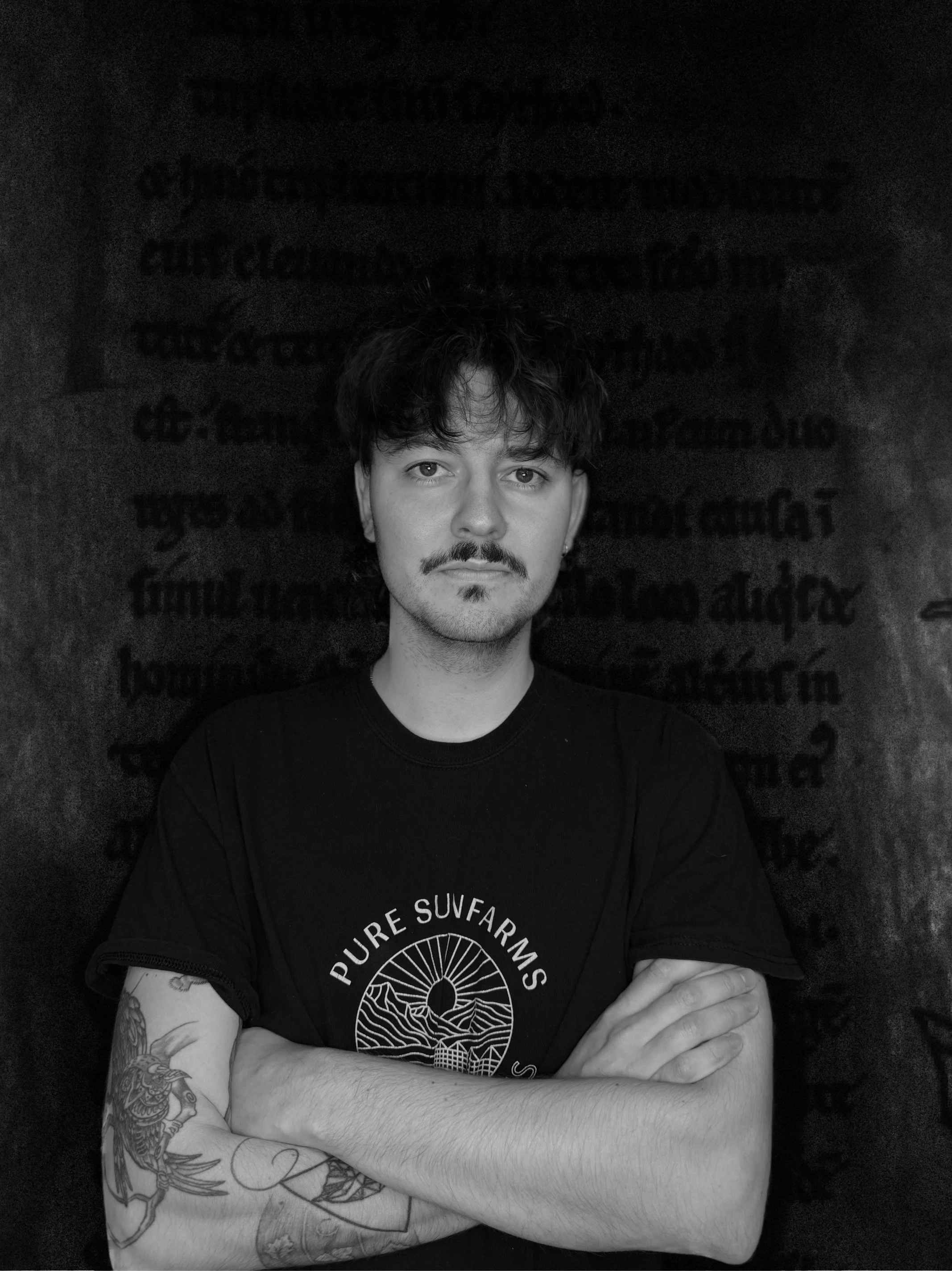 A young man with dark wavy hair and a mustache, wearing a black T-shirt, is standing with arms crossed in front of a dark background with faint text. He has tattoos on his right arm.