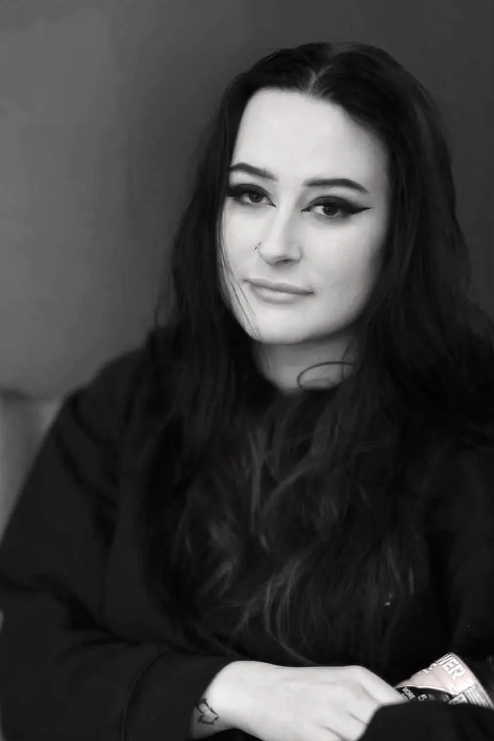 Black and white photo of a woman with dark hair, one eye winking, and a slight smile, wearing dark clothing and a nose ring.