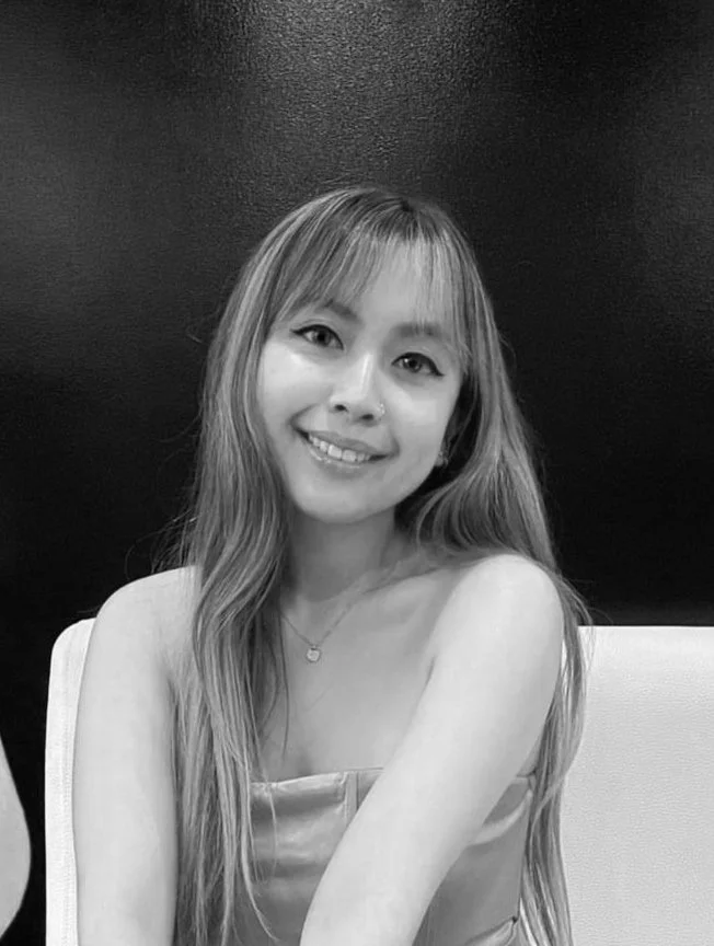 A woman with long hair smiling, sitting against a black background, in black and white.