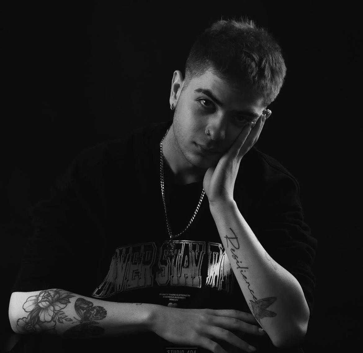 Black and white portrait of a young man with short hair and tattoos, posing with one hand against his face and the other arm resting on a surface, wearing a chain necklace and a dark shirt.